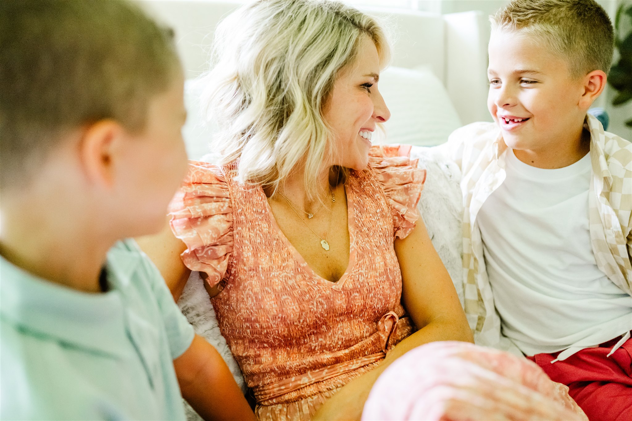 a young boy in a white shirt laughs with mom in a pink dress on a bed