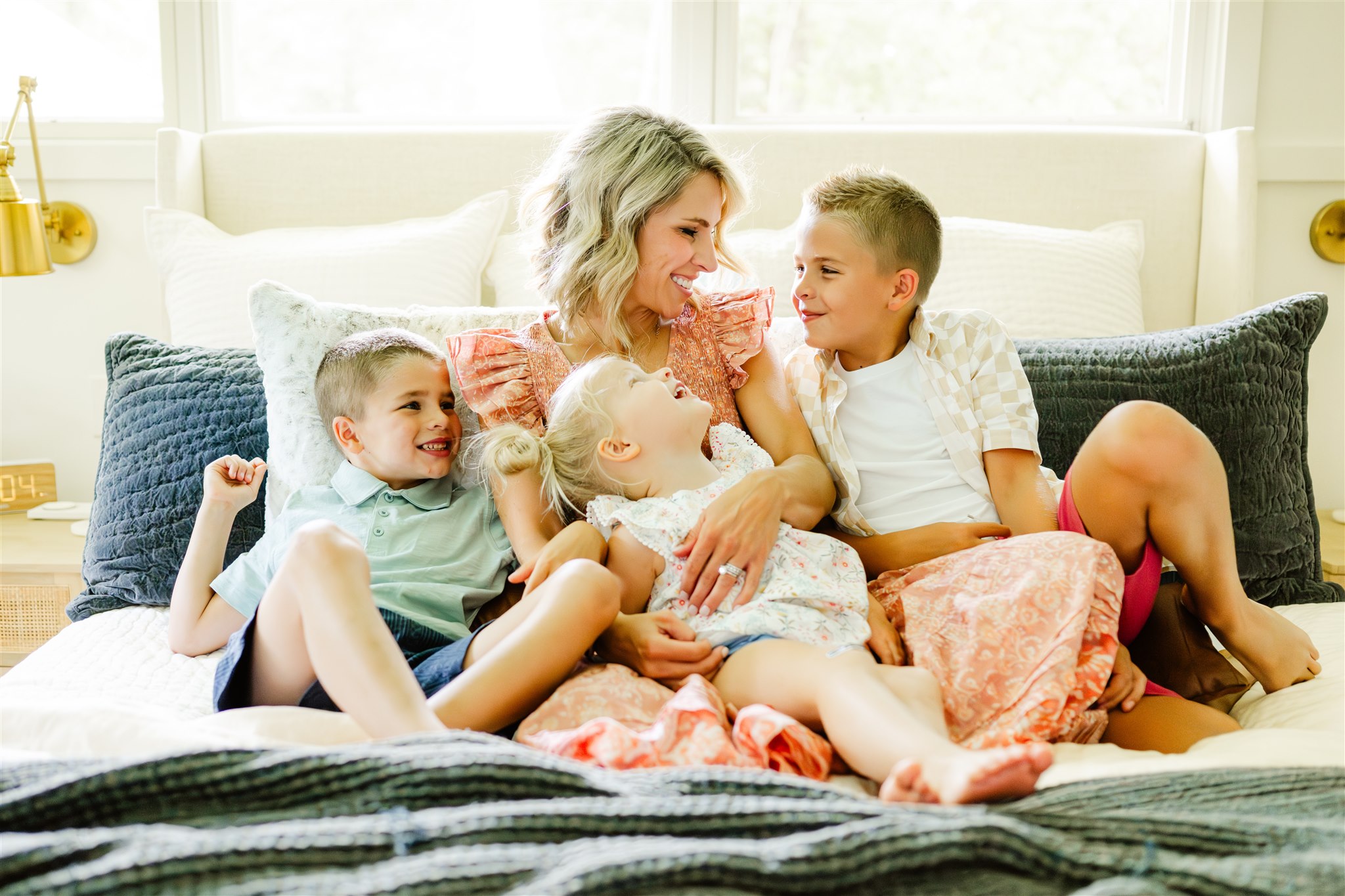 a smiling mom in a dress sits on a bed cuddling and laughing with her three toddlers after some family counseling in naperville