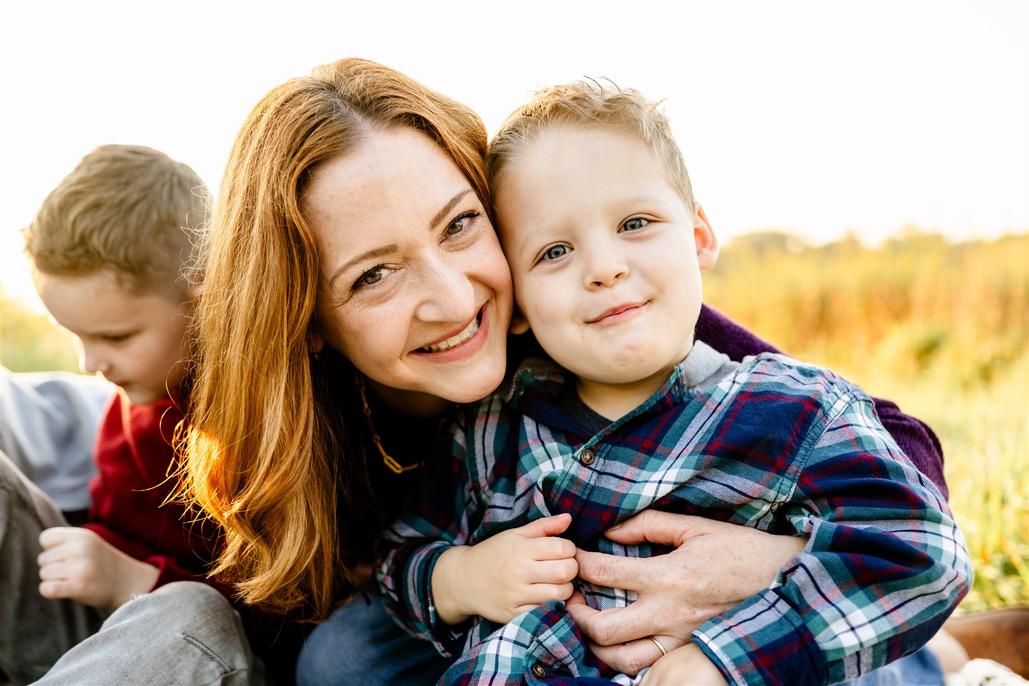 A smiling mom with red hair hugs her toddler son in a plaid shirt while sitting in a park field at sunset