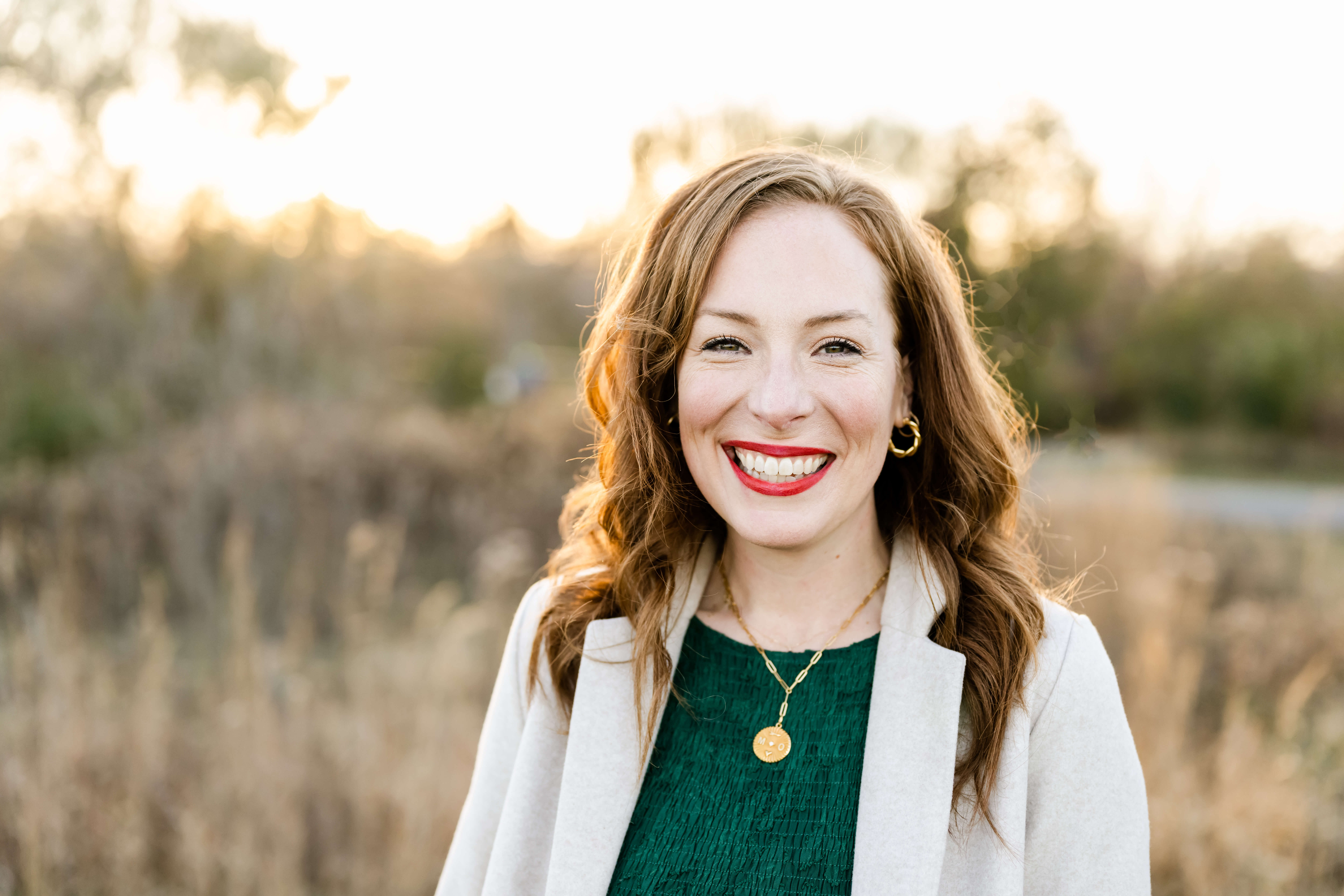 A closeup of a smiling woman with red hair in a green dress and white coat