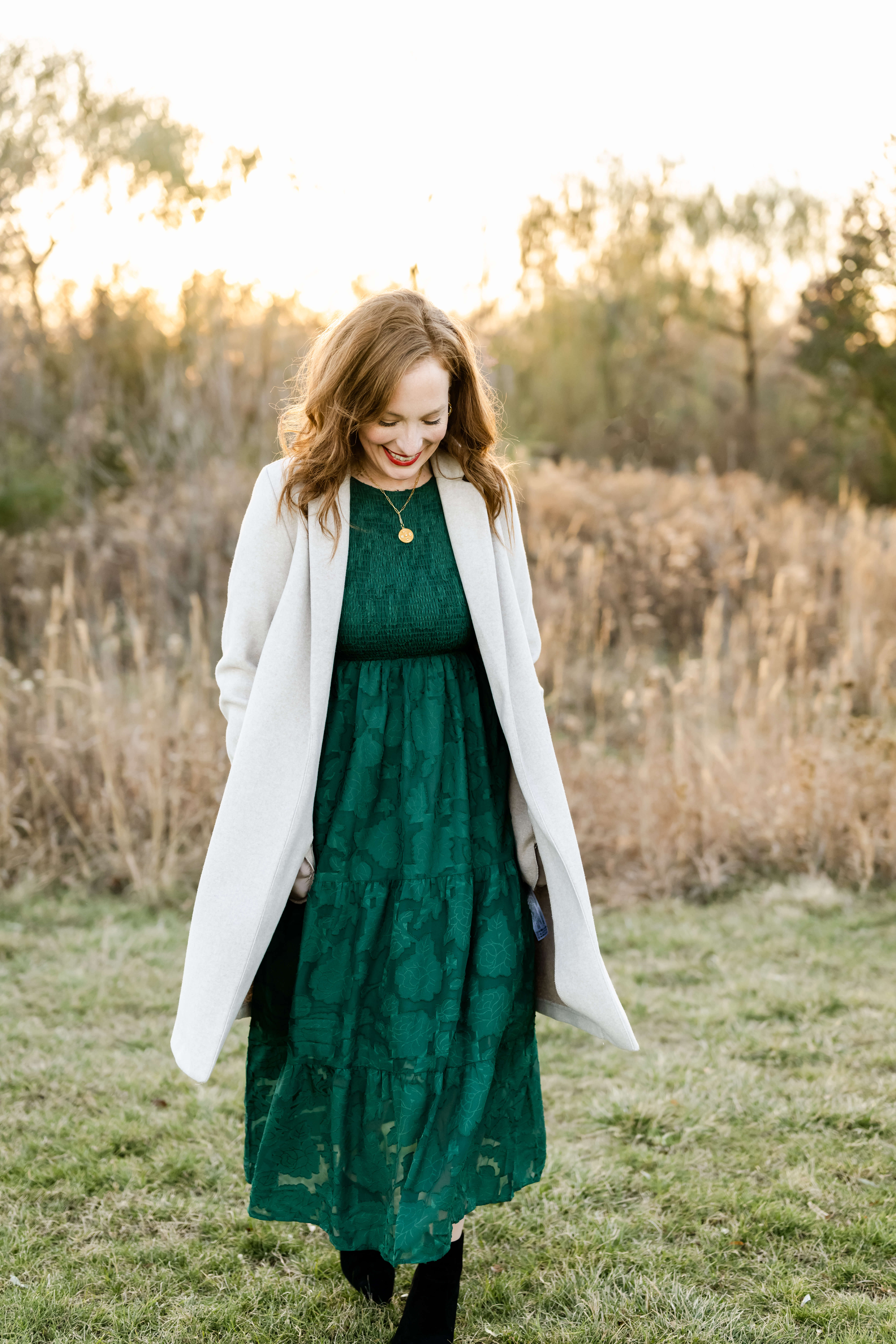 A smiling woman in a green dress and white sweater walking in a field with tall golden grass at sunset before attending Advent by Candlelight