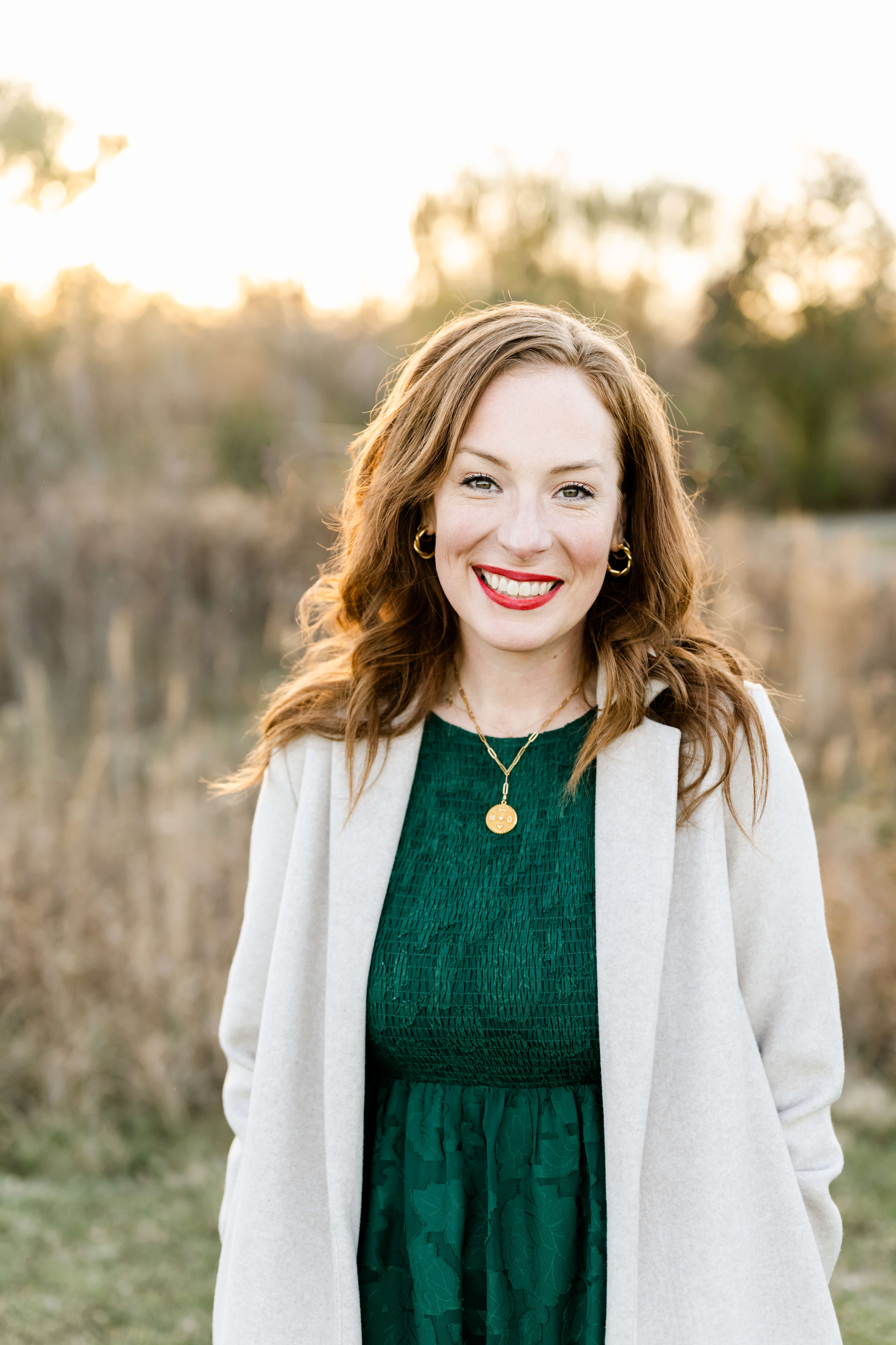 A woman with red hair stands in a field at sunset in a green dress and white sweater smiling before attending Advent by Candlelight