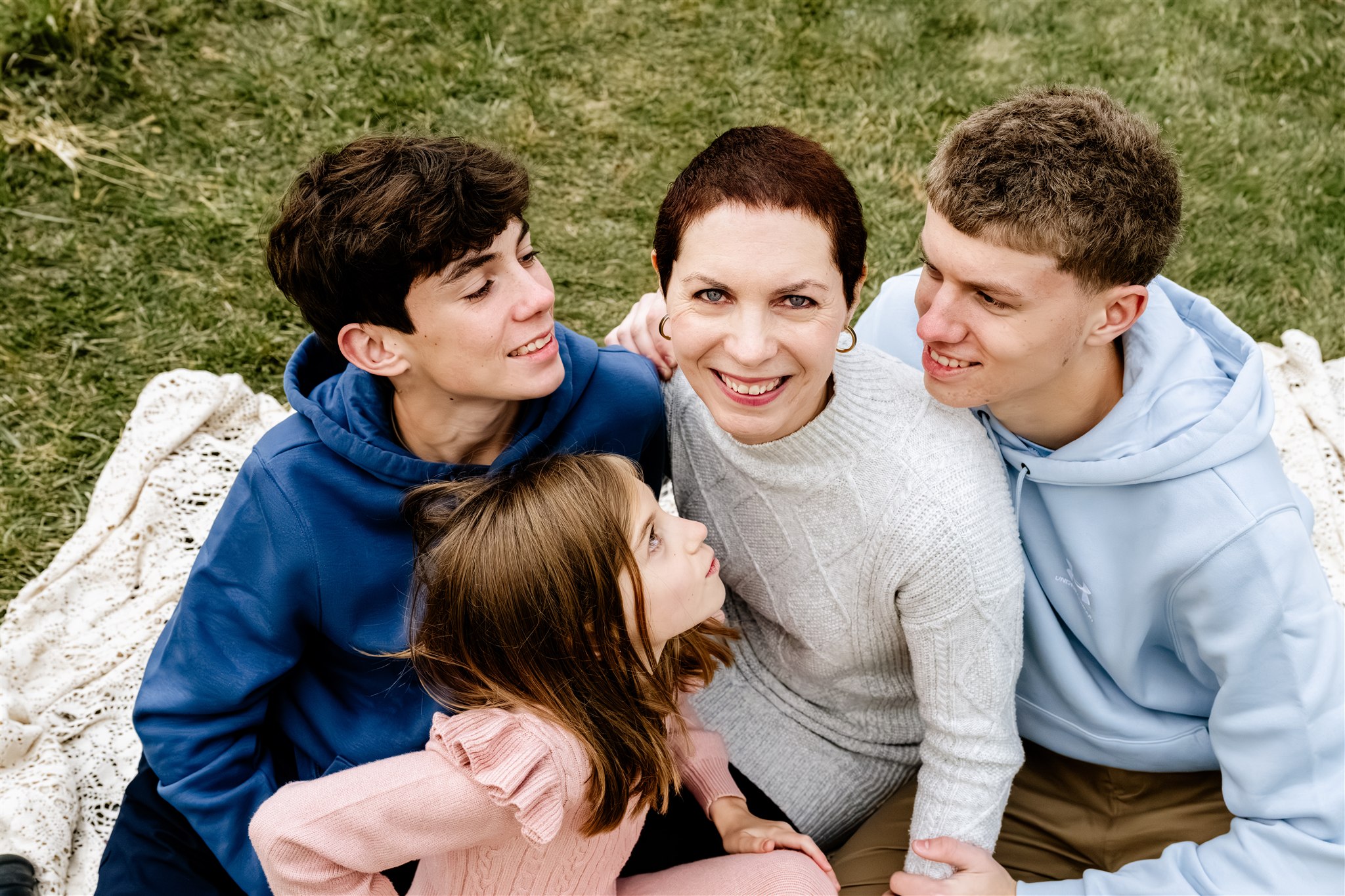 A smiling mother sits on a picnic blanket being hugged by her two teen sons and young daughter