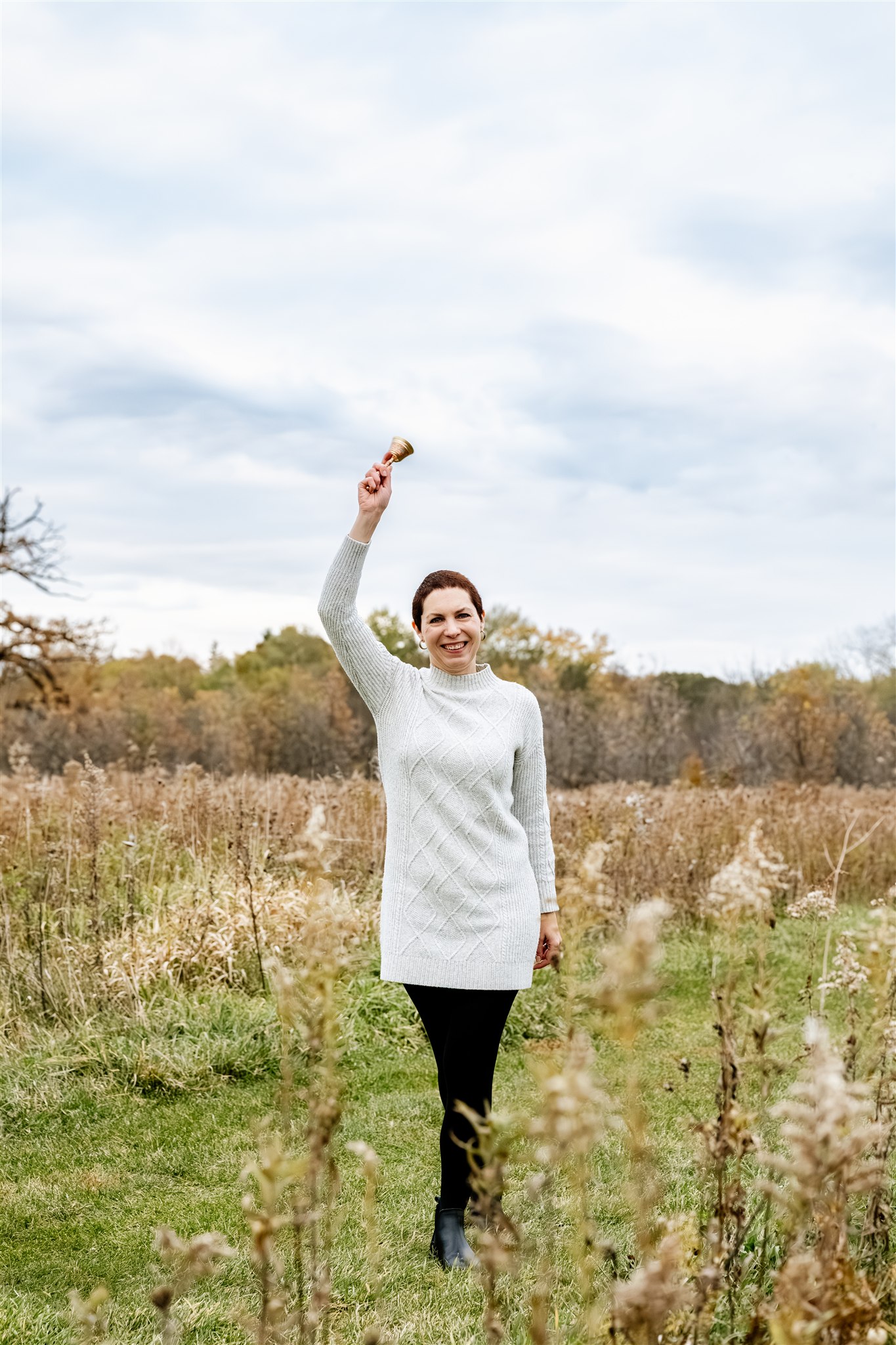 A woman in a white sweater stands in a field of tall grass while ringing a bell above her head thanks to Cancer Support Center in Chicago