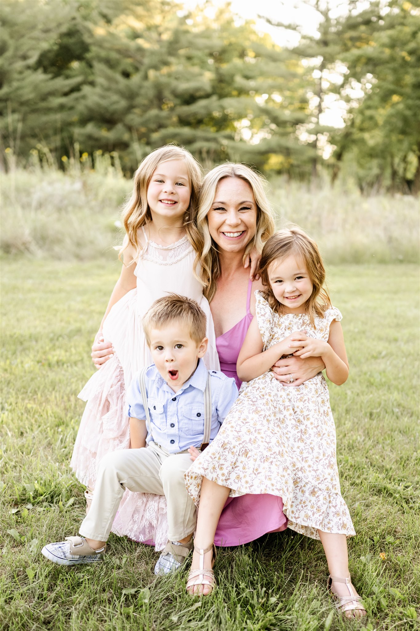 A smiling mom in a pink dress hugs her three toddlers in her lap in a field at sunset