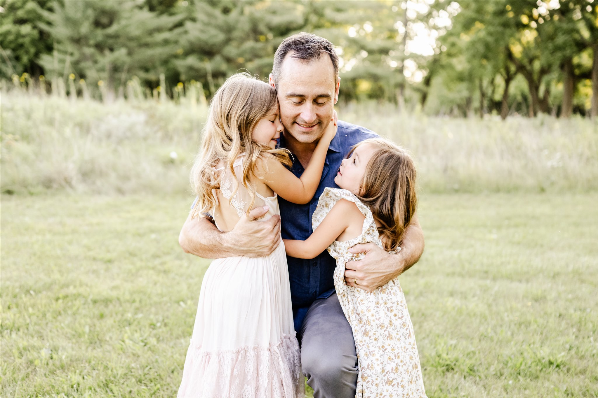 A father in a blue shirt hugs his two toddler daughters in white dresses in a field at sunset after some family counseling in Chicago