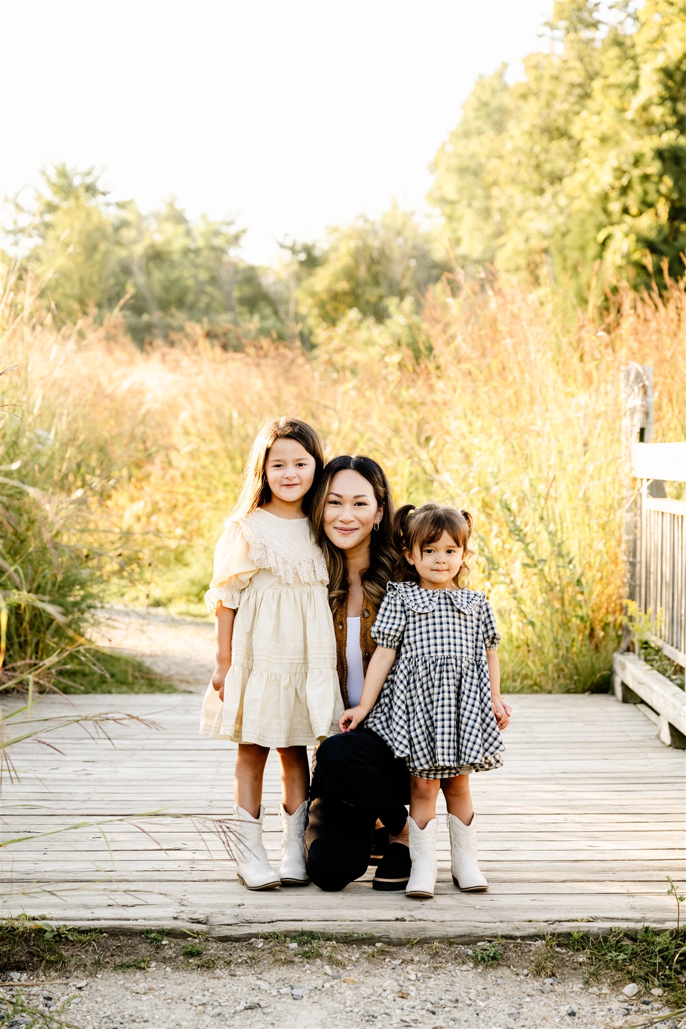 A smiling mother kneels on a boardwalk with her two toddler daughter in black and white dresses