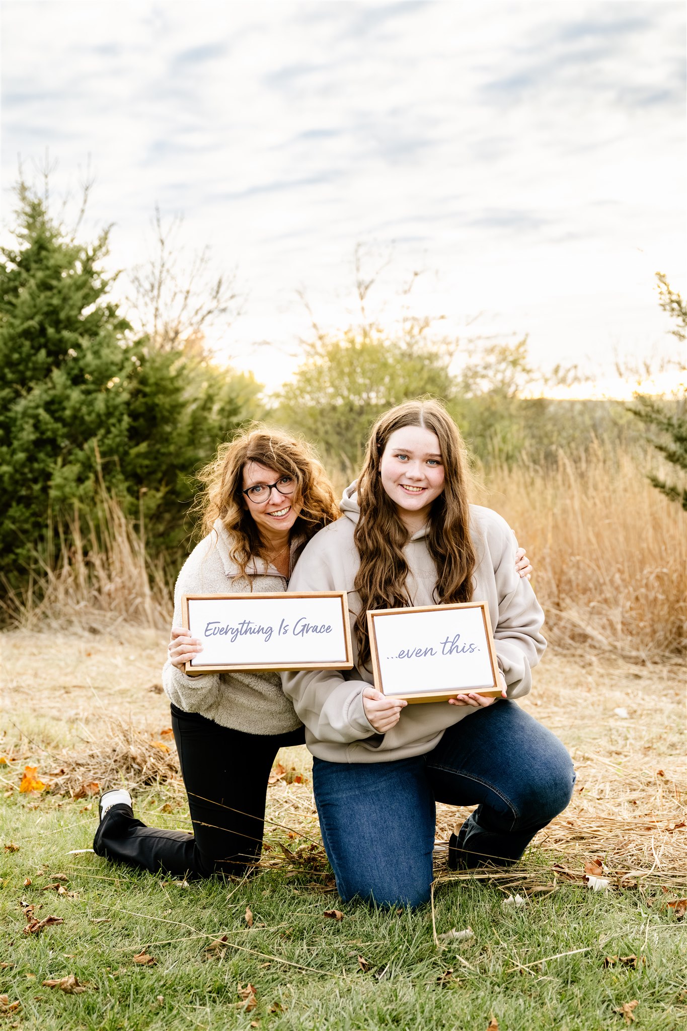 A happy mom hugs her daughter as they kneel holding signs in a field in tan sweaters
