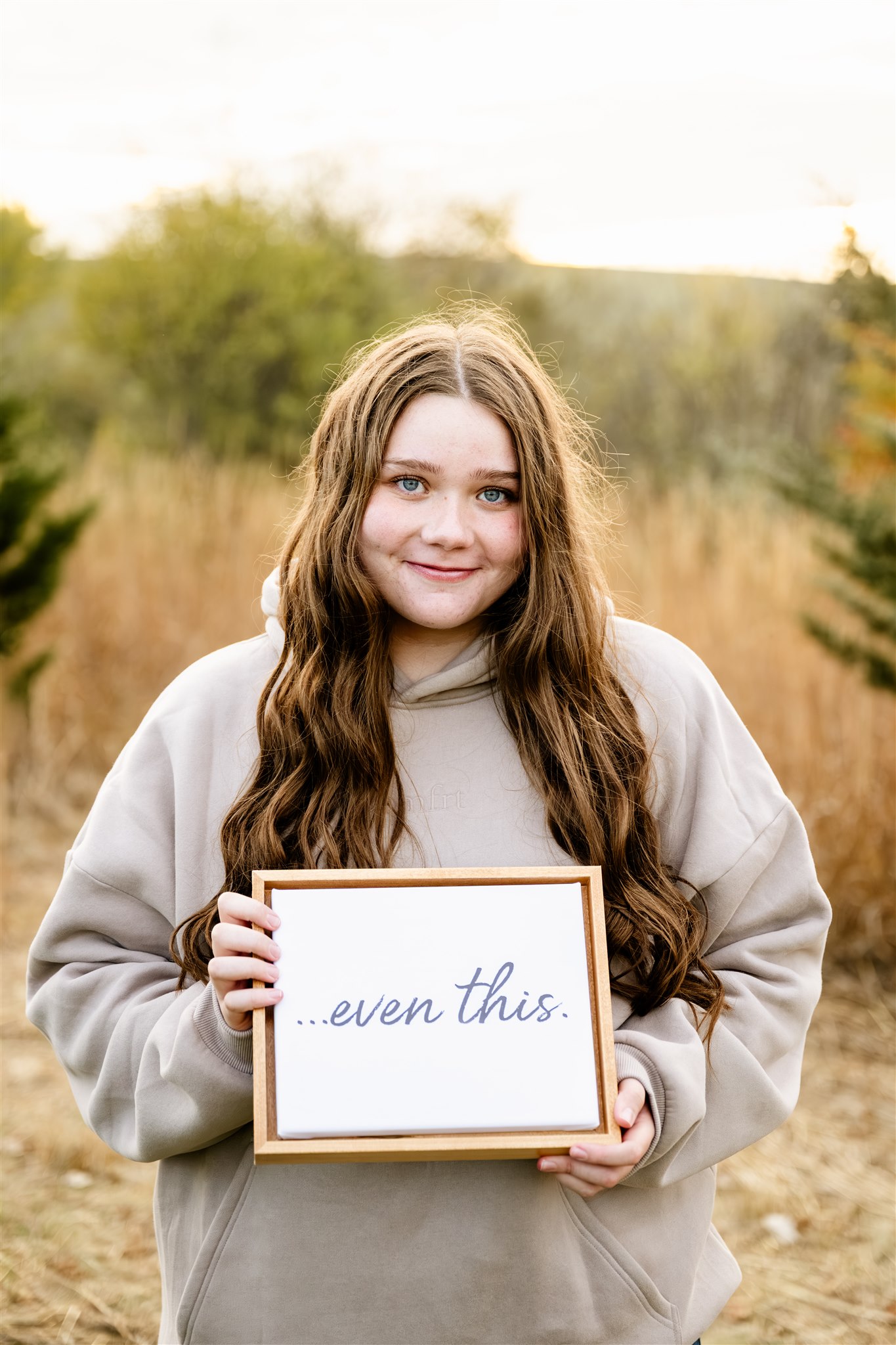 A woman in a tan hoodie holds a sign saying "...even this." in a field of tall golden grass at sunset while smiling thanks to Loyola Hospital Cancer Center
