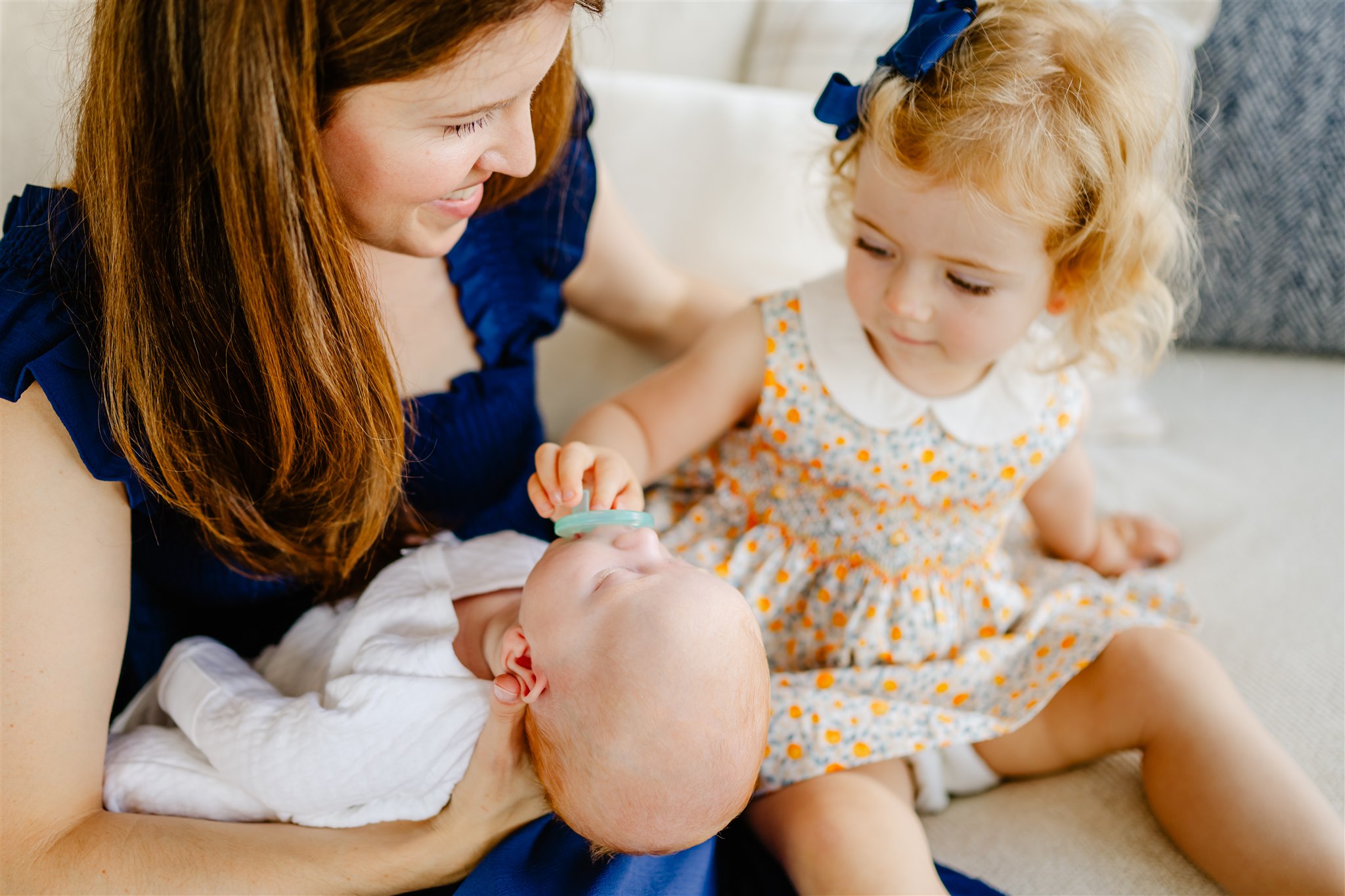 A toddler girl in a yellow dress helps hold her newborn siblings binky while laying in mom's arms on a couch
