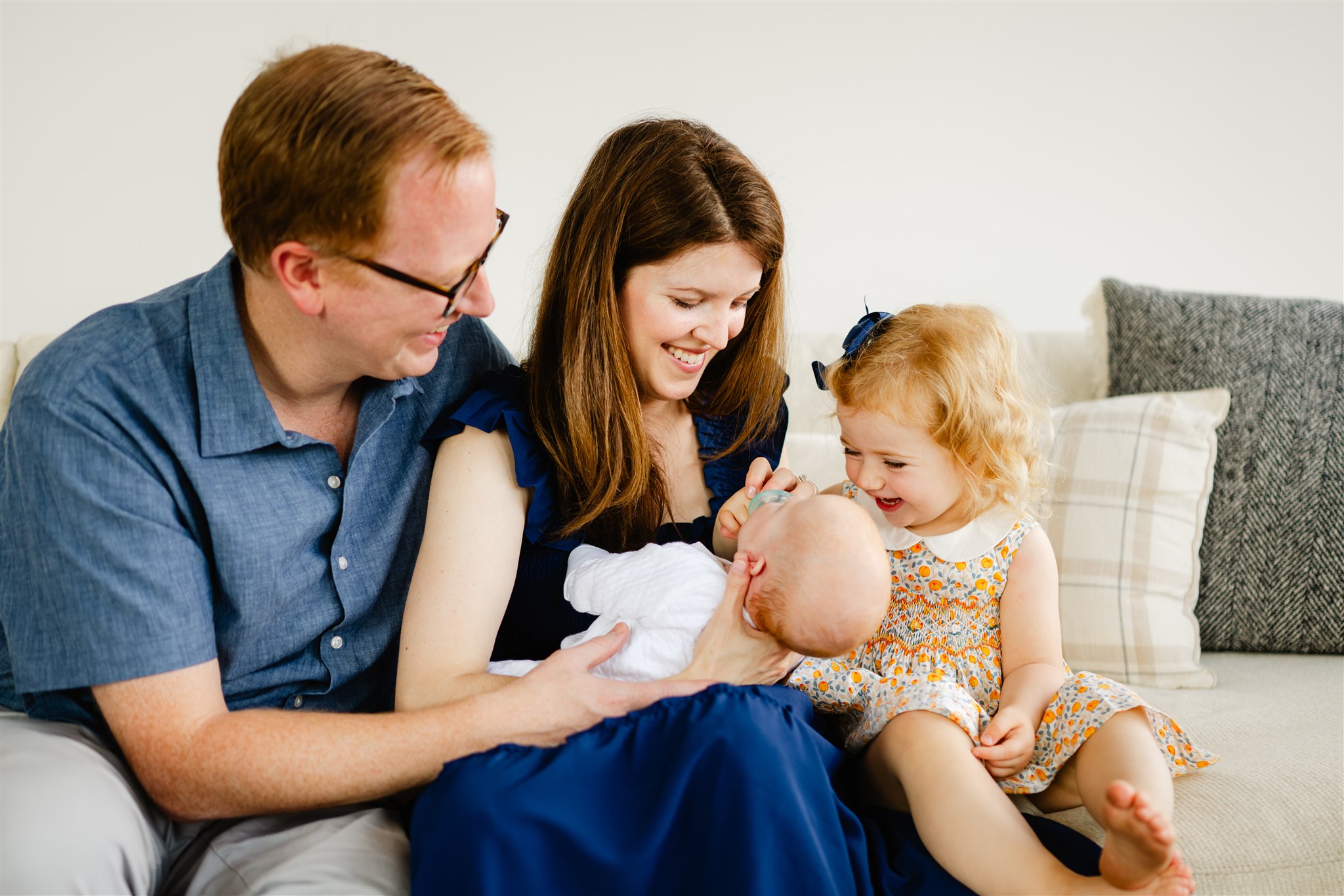 happy mom and dad in blue outfits play with their newborn and toddler while sitting on a couch after some parenting classes in chicago