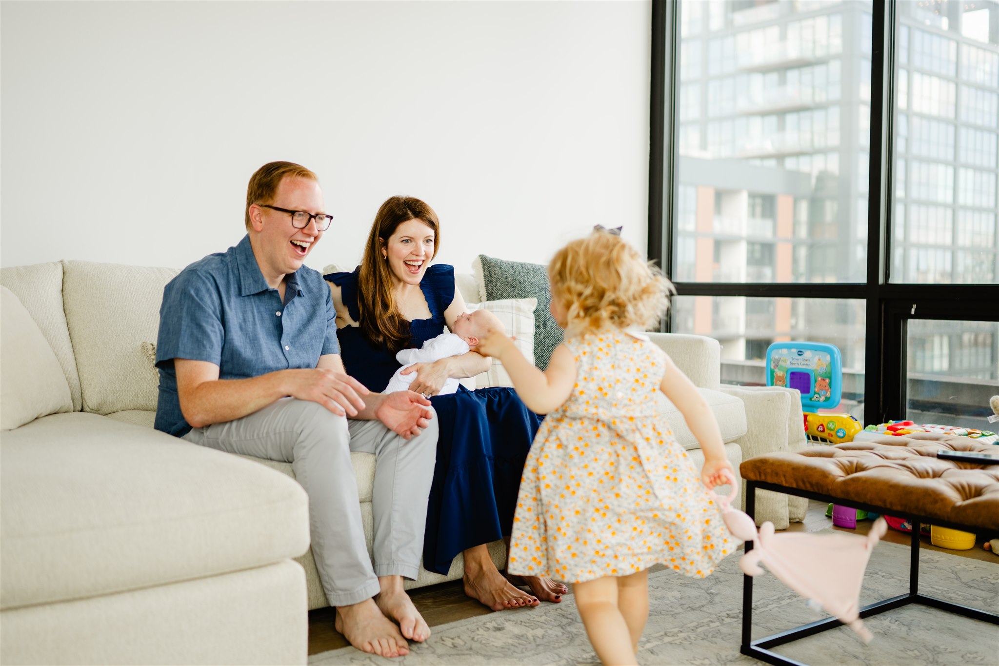 a mom and dad on a couch in blue outfits holding a newborn happily welcome their toddler walking towards them in a dress after some parenting classes in chicago