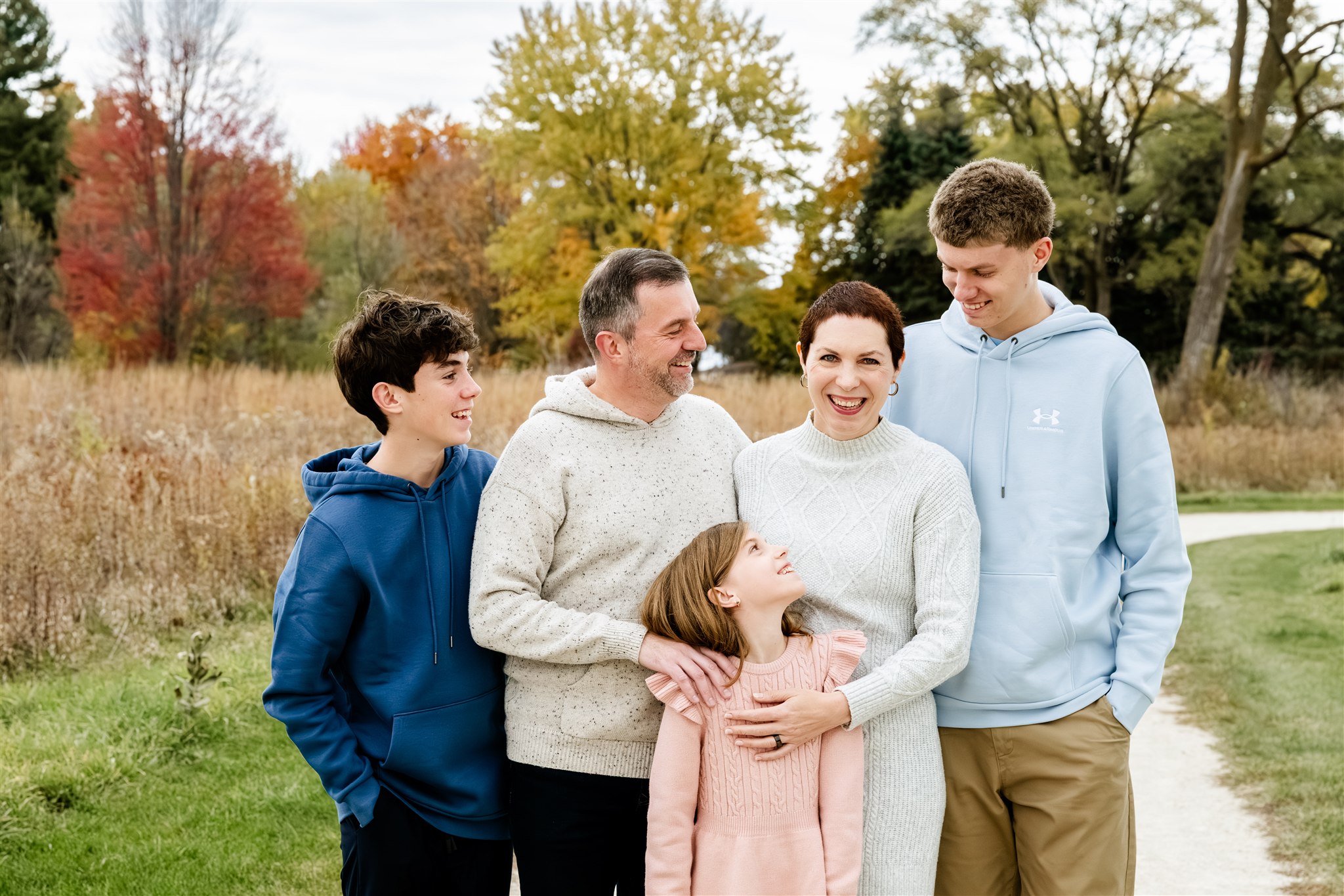 A smiling mother in a white sweater dress as her husband and three children smile at her in a park trail in fall