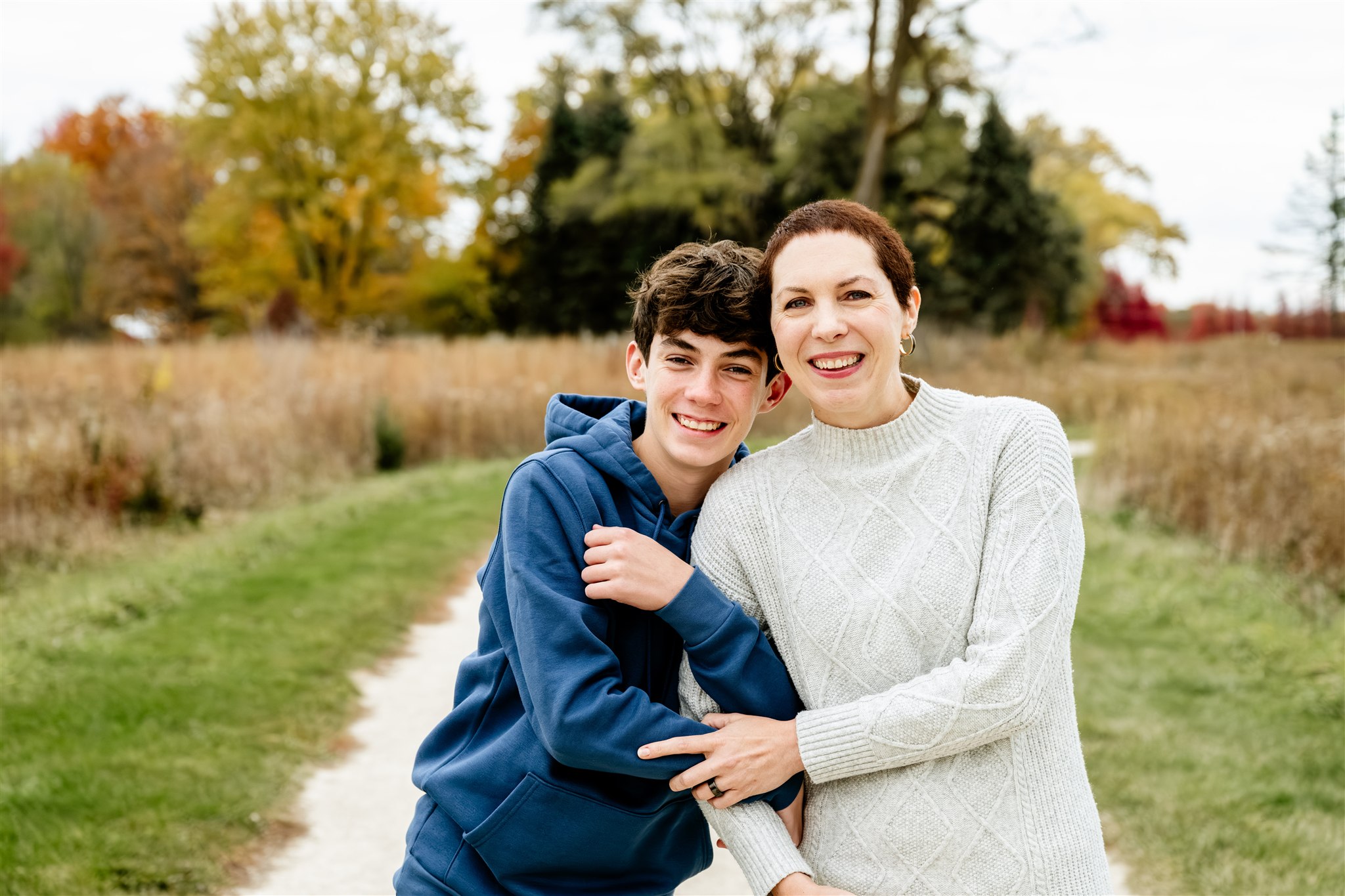 A smiling teen boy in a blue hoody hugs on the arm of his mother in a white sweater while walking a park in fall after visiting Wellness House in Hinsdale, IL