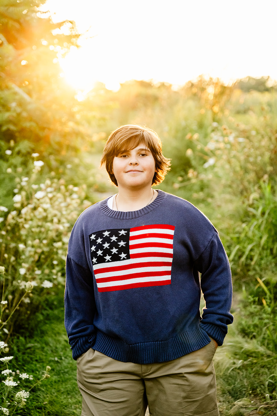 A smiling high school grad stands with hands in their pockets smiling in a tight park trail at sunset