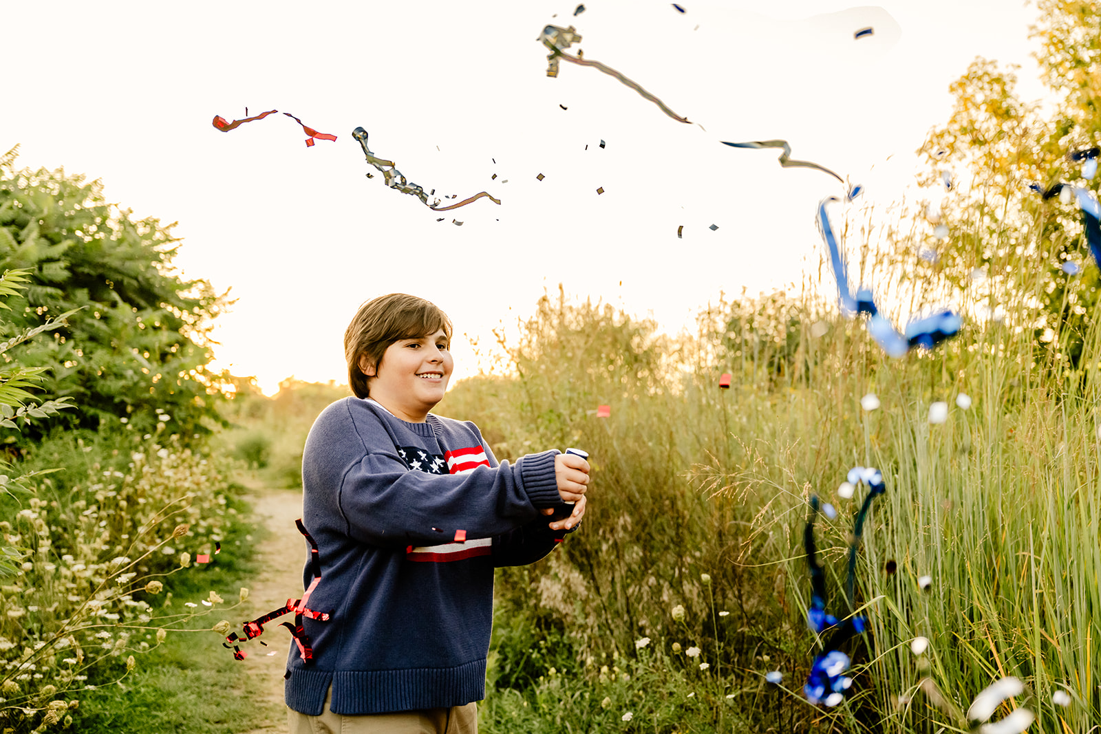 A high school senior in a blue american flag sweater pops a streamer cannon in a park trail at sunset thanks to ACT prep in Naperville