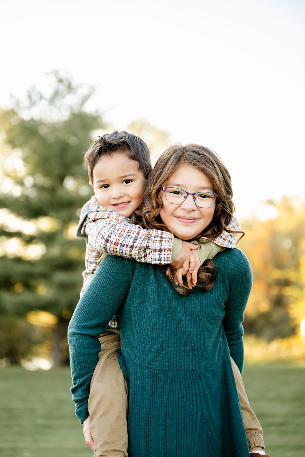 A young girl in a green dress carries her younger brother on her back in a park at sunset