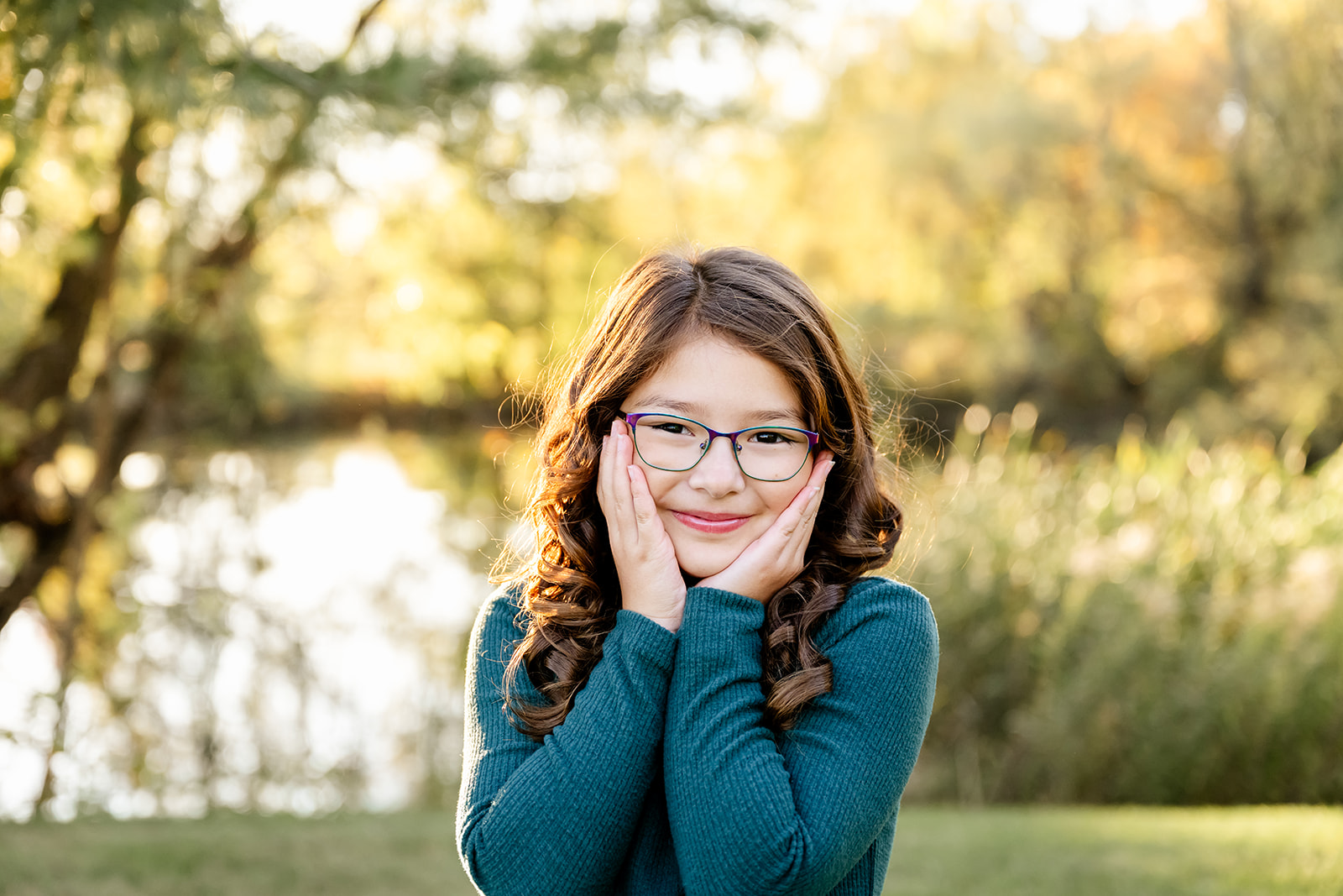 A young girl holds her hands on her cheeks in a blue sweater by a pond at sunset after visiting catholic schools in Naperville