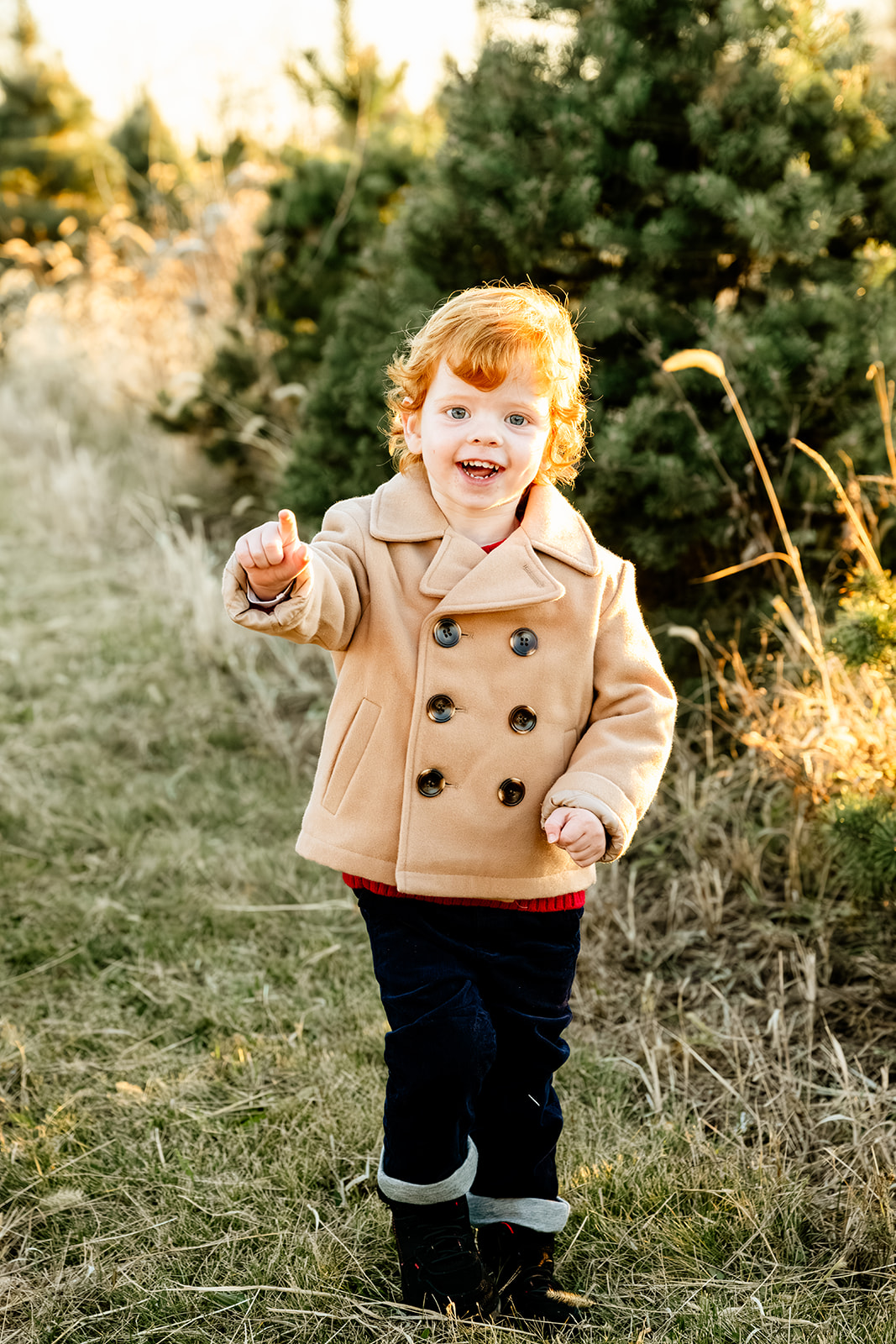 A young toddler in a tan coat and black pants points with a smile in a christmas tree farm before visiting indoor playgrounds in Naperville
