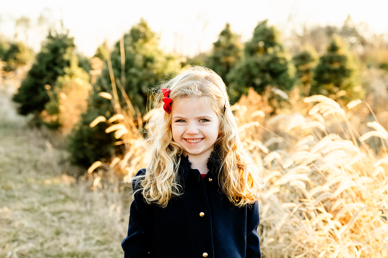 A smiling toddler girl in a black coat explores tall golden grass at a christmas tree farm before visiting indoor playgrounds in Naperville