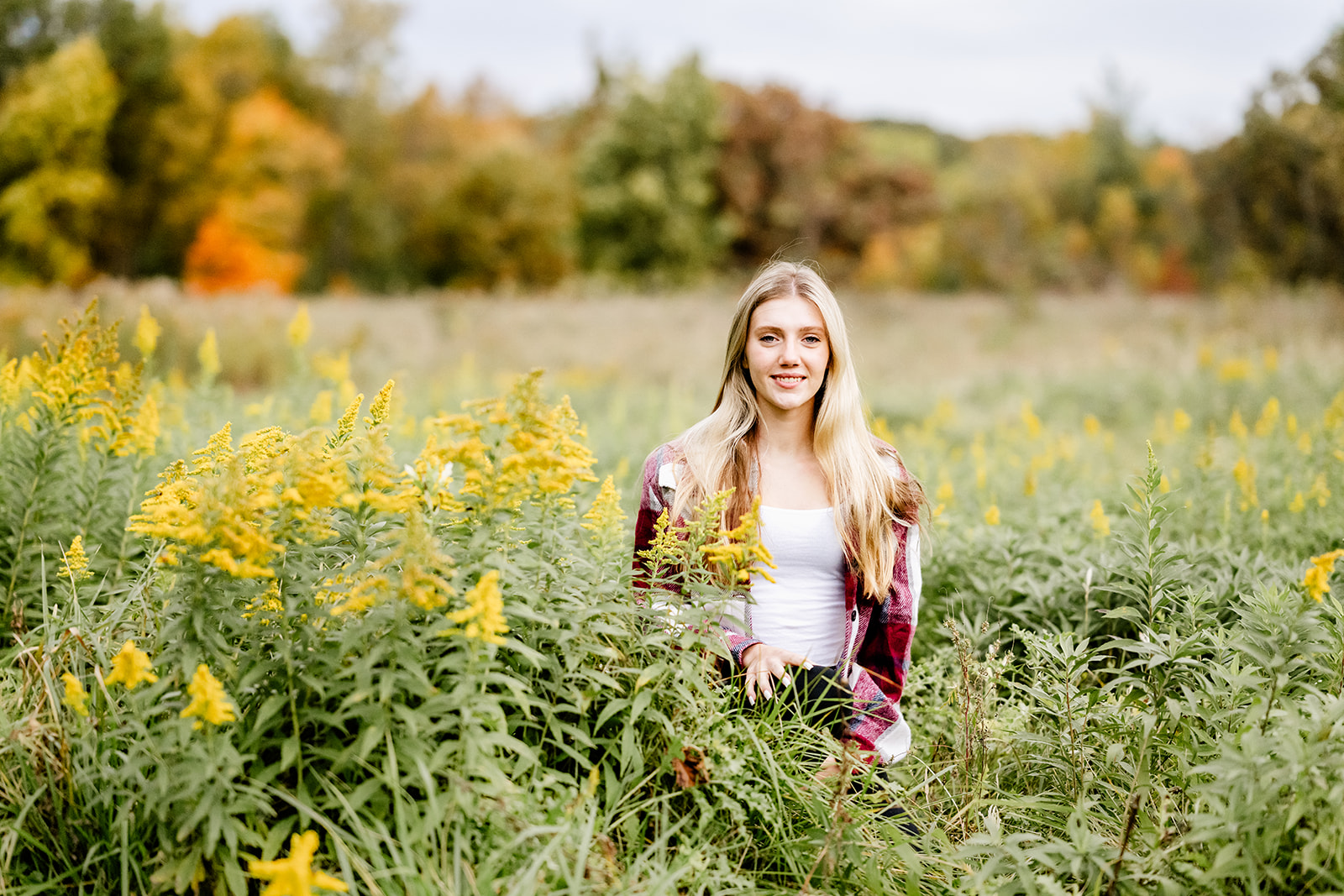 A high school senior kneels in tall gold and yellow grasses in a field in a red sweater and white blouse after loving private catholic schools in Chicago