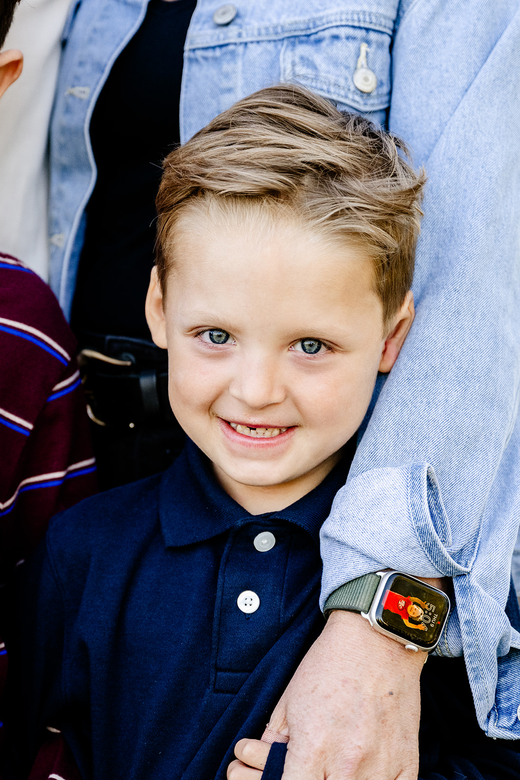 A young boy stands against mom in a blue polo while holding her hand