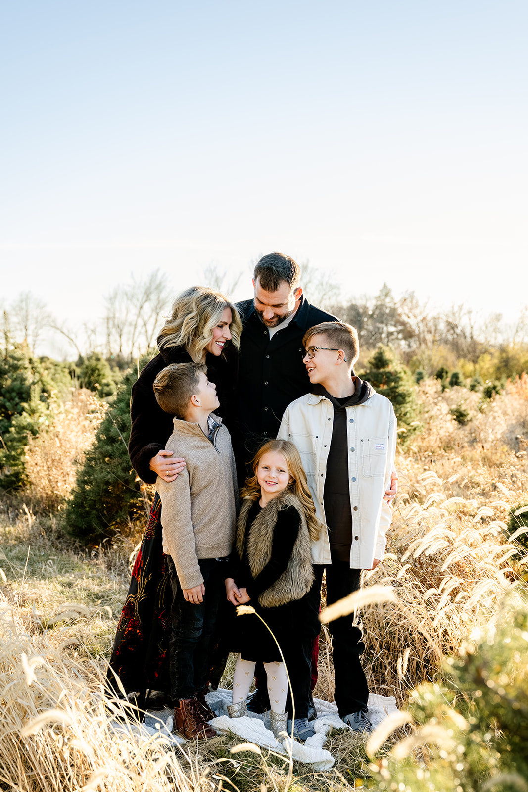 Happy mom and dad stand in a christmas tree farm hugging their three young children at sunset