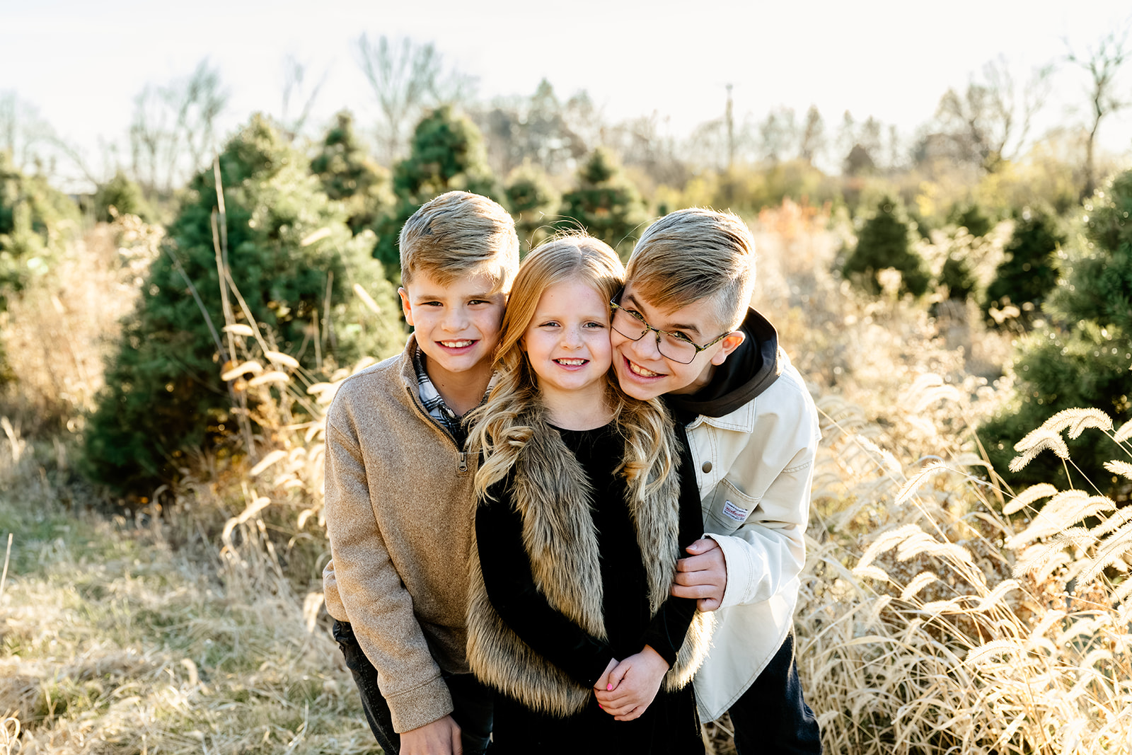 Three young happy siblings hug and smile in a christmas tree farm at sunset after visiting private elementary schools in Naperville