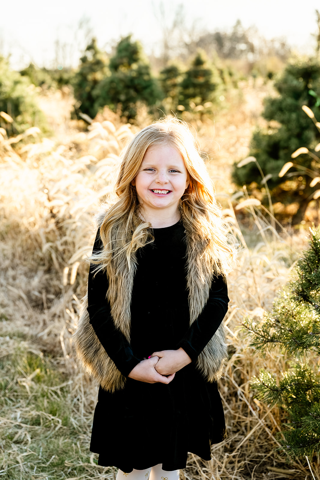 A toddler girl in a black dress and fur vest smiles in a park lawn after exploring private elementary schools in Naperville