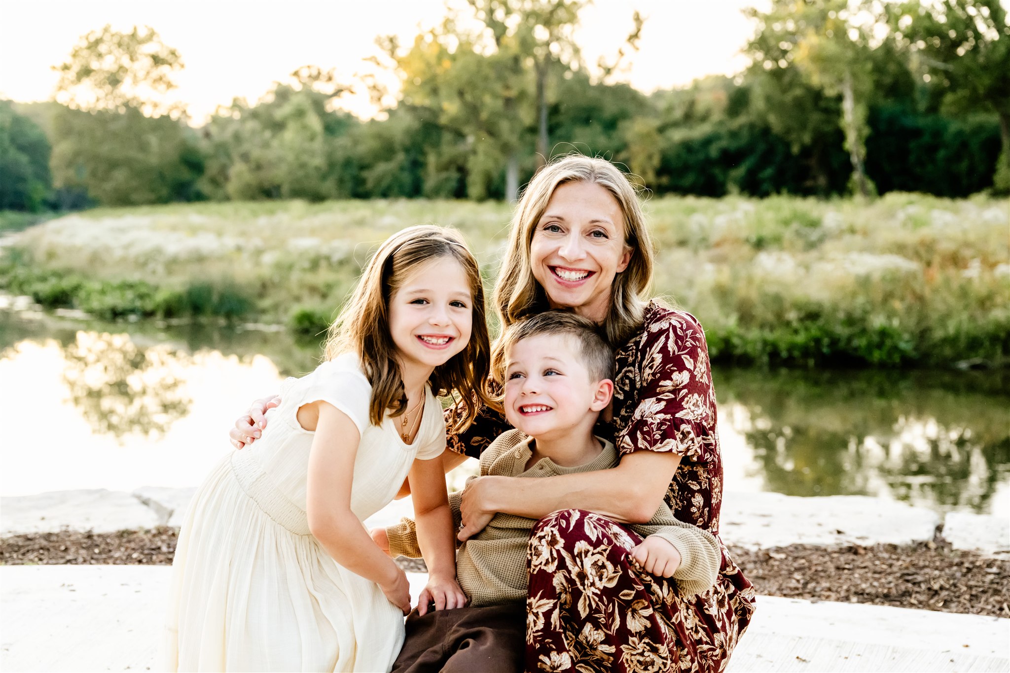 A smiling mom in a maroon floral print dress kneels while hugging her two happy toddlers in a park by a pond at sunset