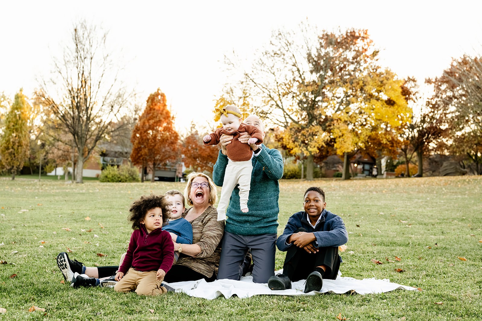 A dad lifts his baby girl up while sitting on a picnic blanket with mom and three older children in a park in fall at sunset before visiting Children's Museums in Naperville