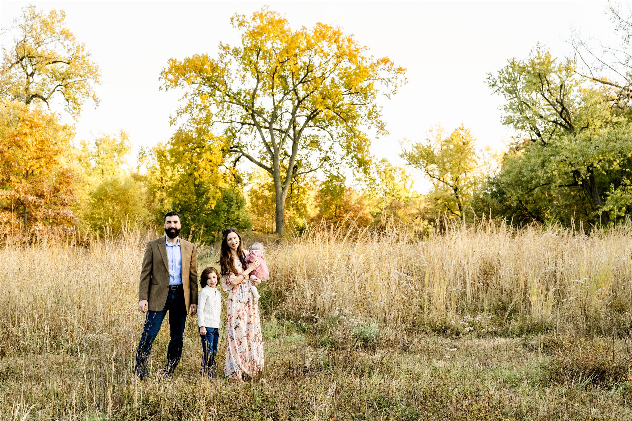 Mom and dad stand in a park with tall golden grass at sunset with their toddler children