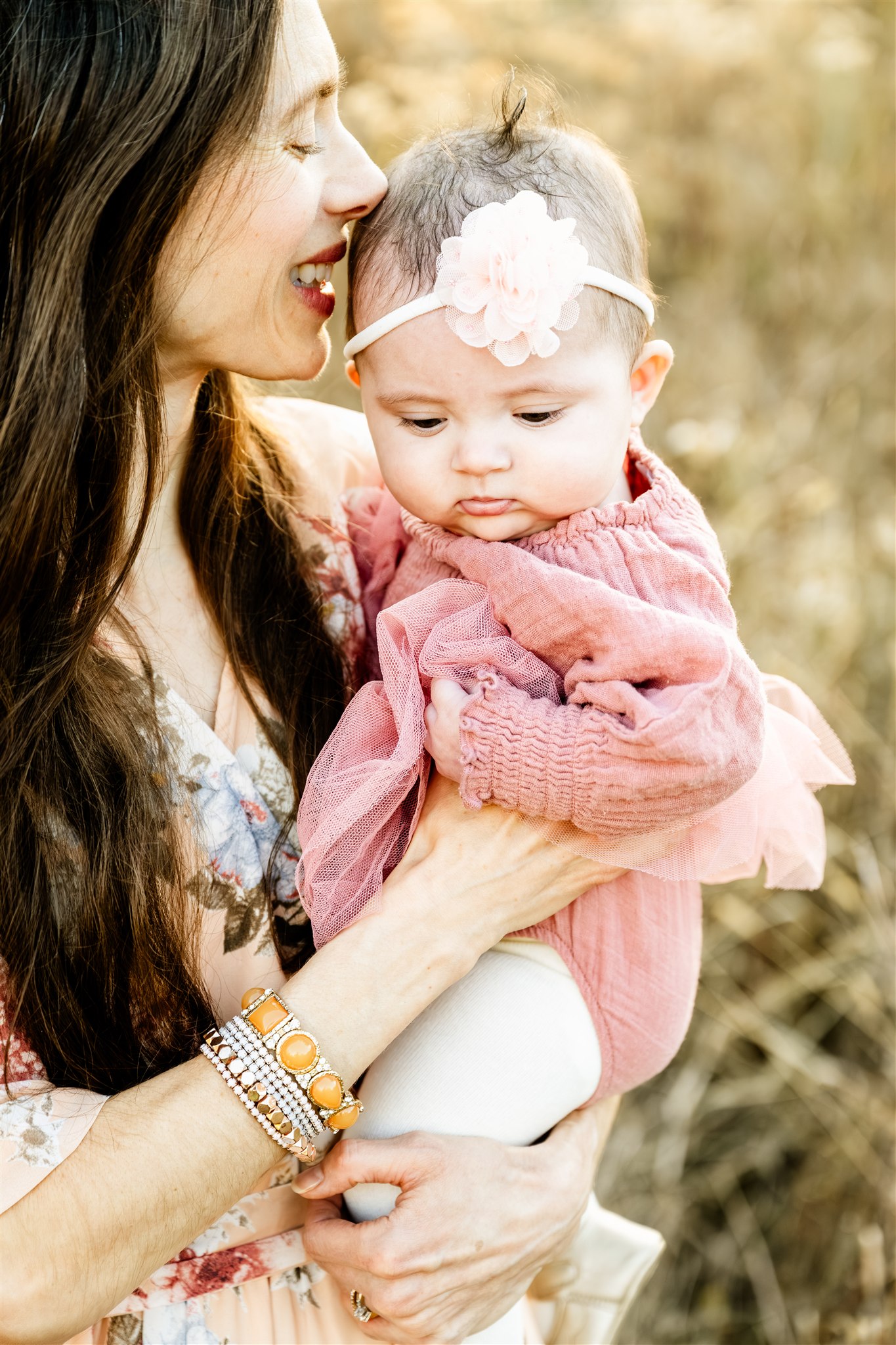 A baby girl sits in mom's arms in a pink onesie as mom kisses her head while standing in tall golden grass at sunset after enjoying pediatric chiropractors in Chicago