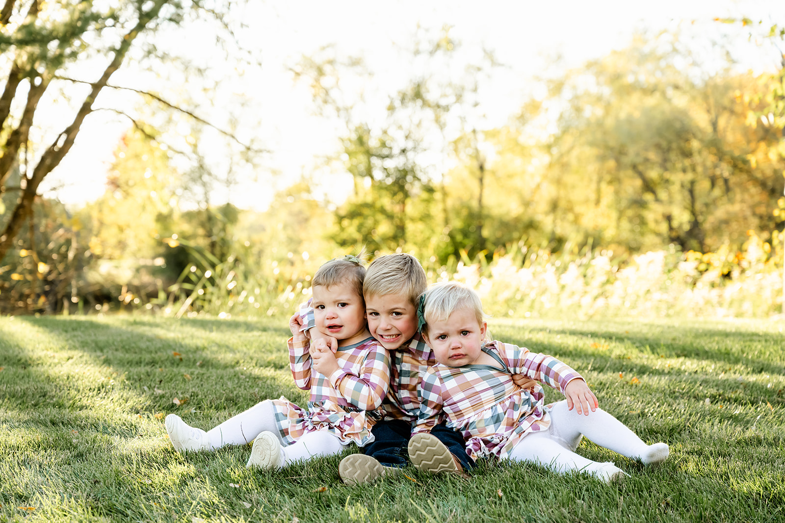 A toddler boy sits hugging his two toddler sisters in matching dresses to his shirt in a park at sunset
