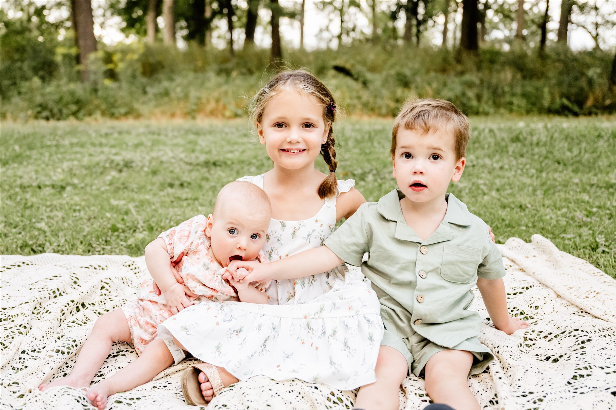 Smiling toddler brother and sister in green and white sit on a picnic blanket in a park with their infant sister in pink