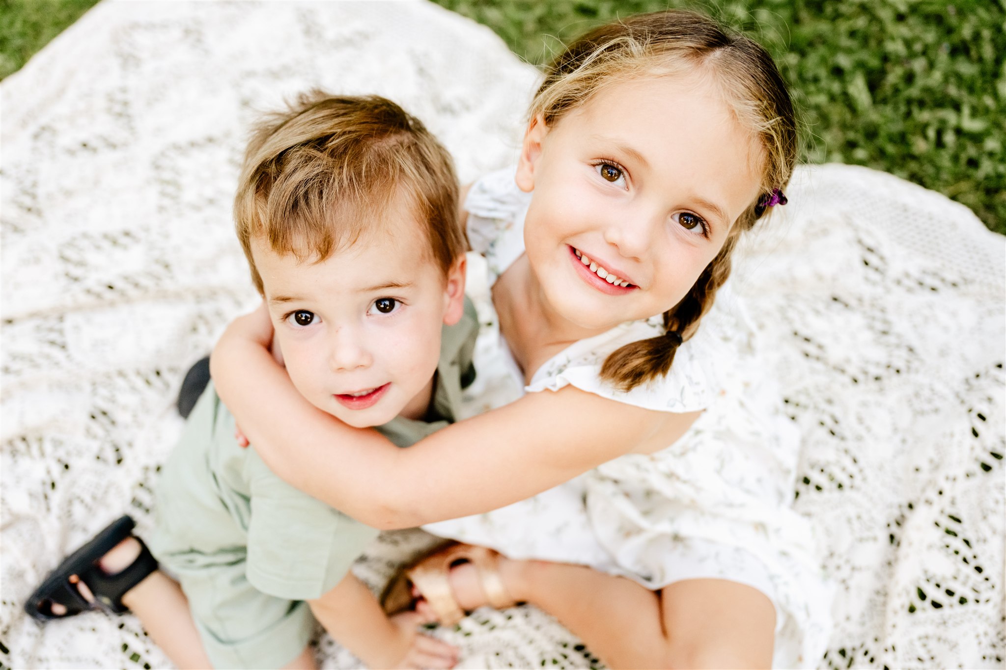 Happy toddler girl in a white dress hugs her younger brother as they sit on a picnic blanket before spring break camps in Naperville