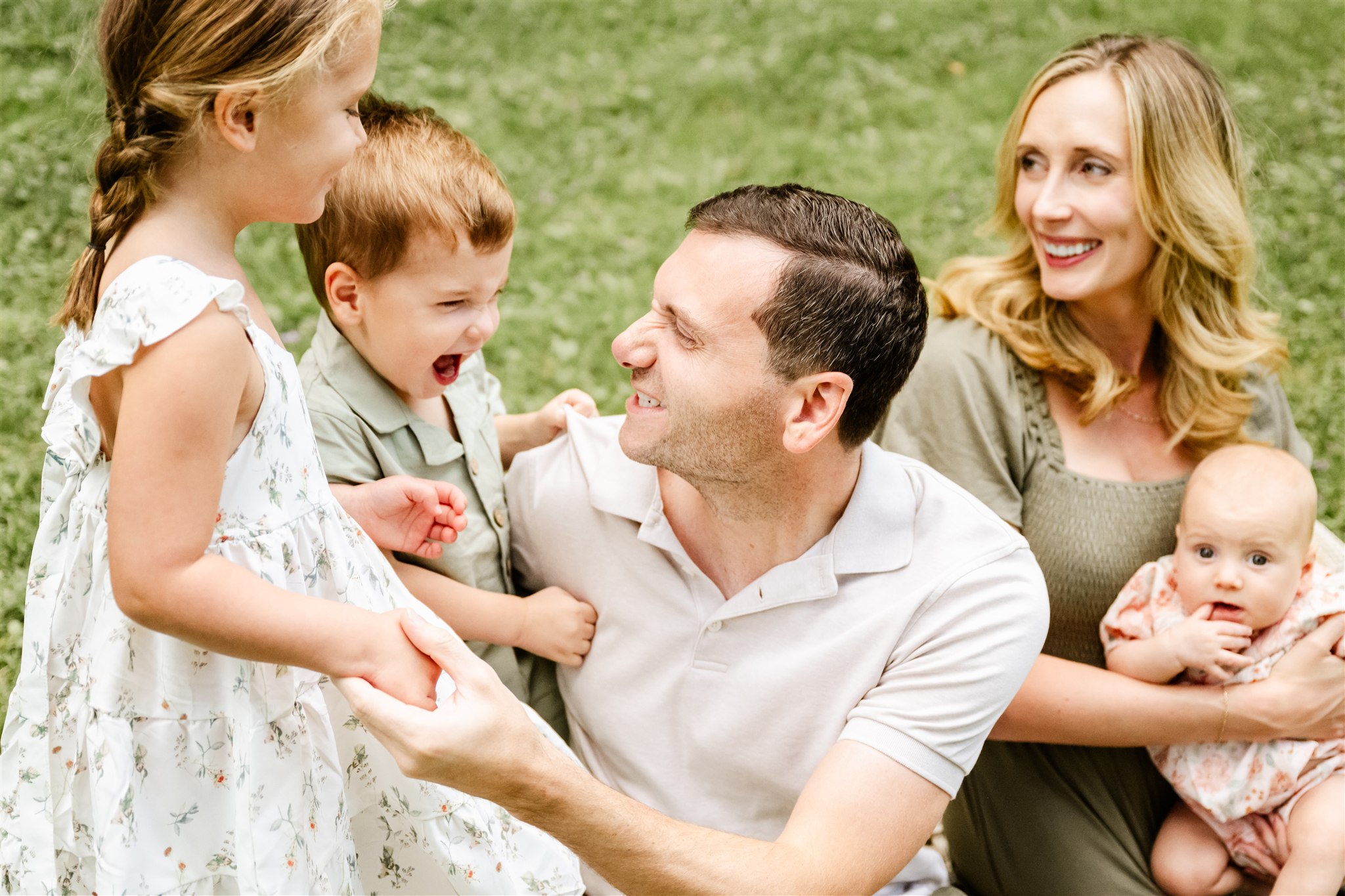 Happy mom and dad sit in a park lawn playing with their two toddlers and infant in mom's lap before some spring break camps in Naperville