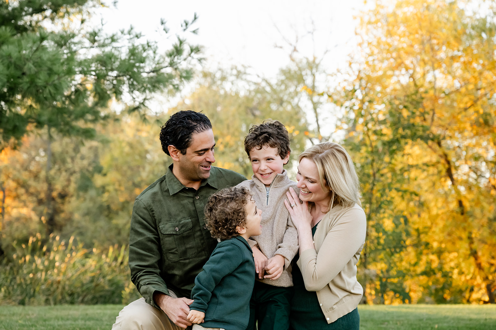 Happy mom and dad play with their two toddler sons in green and tan in a park at sunset