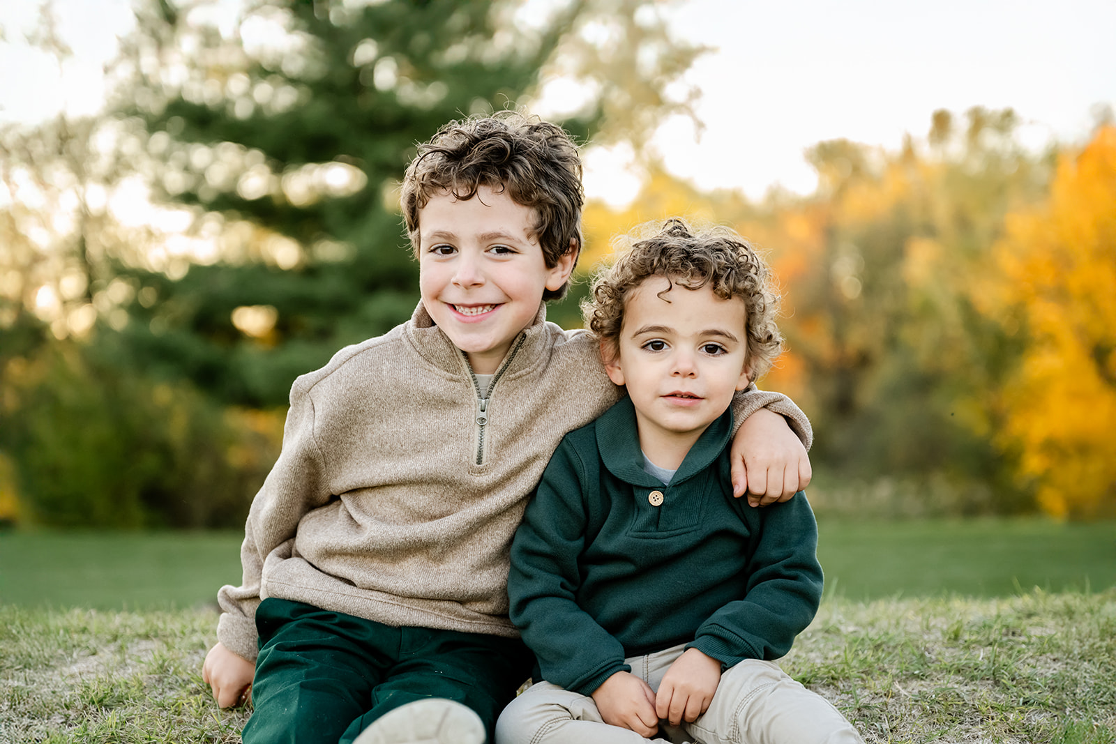 Happy toddler boys in green and tan sweaters sit smiling in a park at sunset after picking summer camps in Naperville