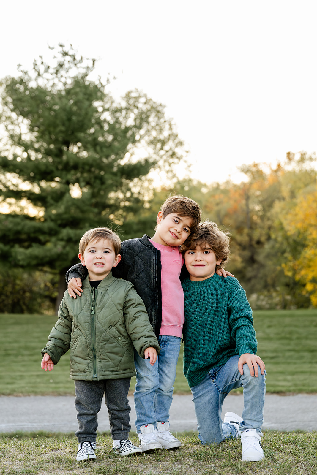 Three toddler brothers stand and kneel ion a park at sunset in sweaters and jackets
