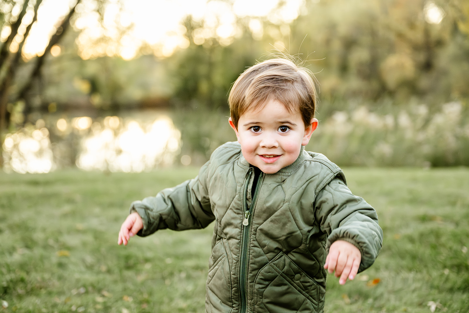 A toddler boy in a green jacket in a park at sunset playing with arms apart after visiting toy stores in Chicago