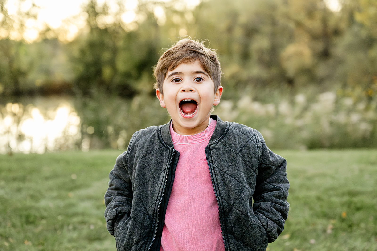 A toddler boy in a black jacket and pink shirt yells with mouth open in a park by a pond after visiting toy stores in Chicago