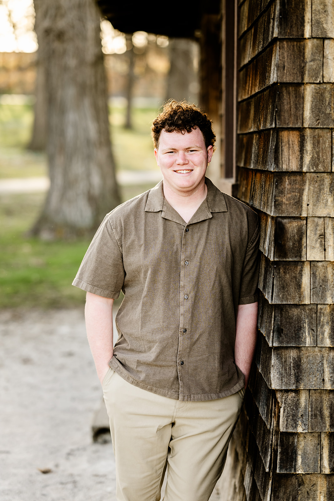 A teenage boy in high school wearing a brown shirt and khakis leans against a rustic building in a park at sunset after finding excellent tuxedo rental in Naperville