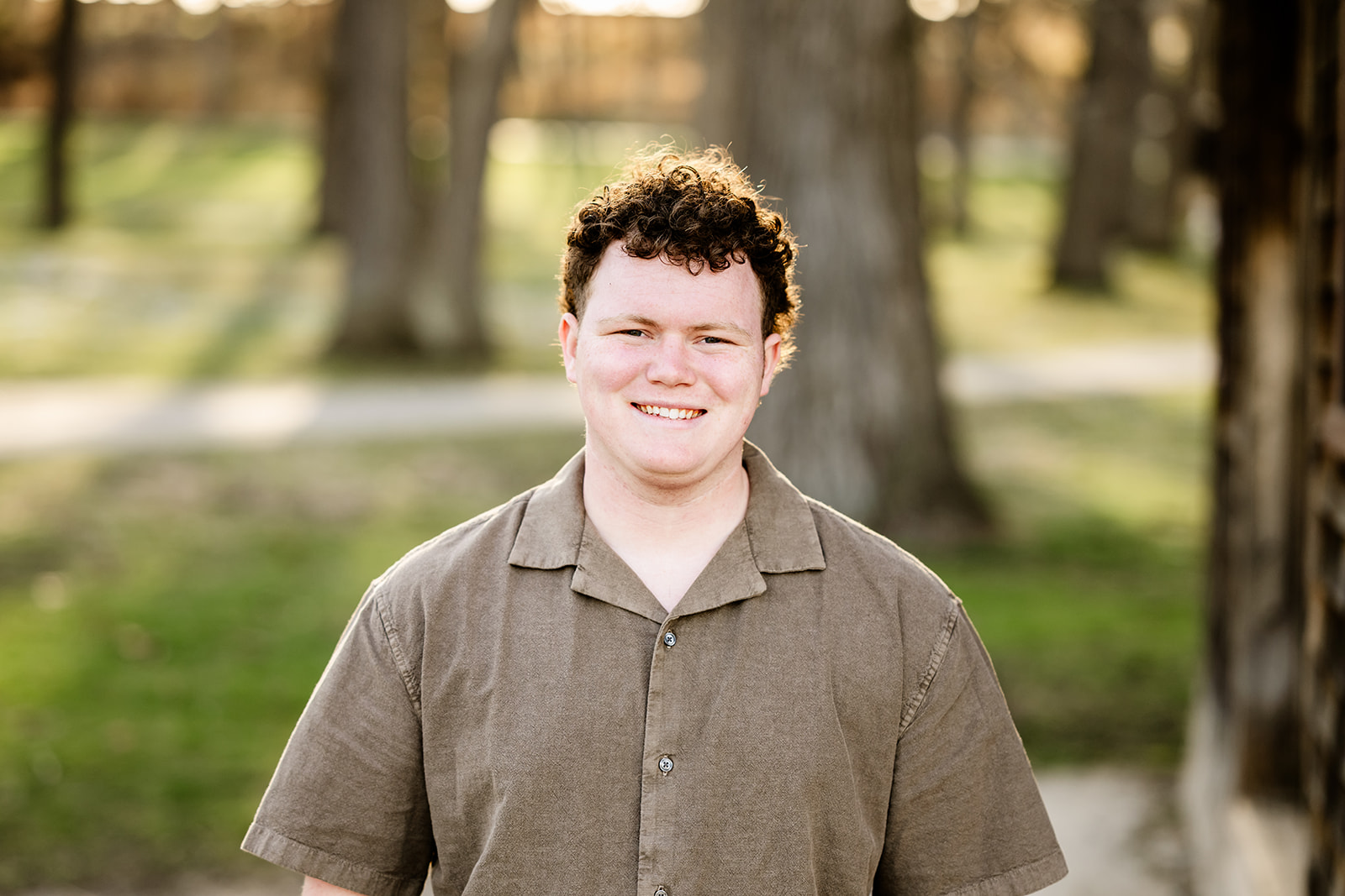 A high school senior in a brown shirt smiles while standing in a park at sunset after enjoying tuxedo rental in Naperville