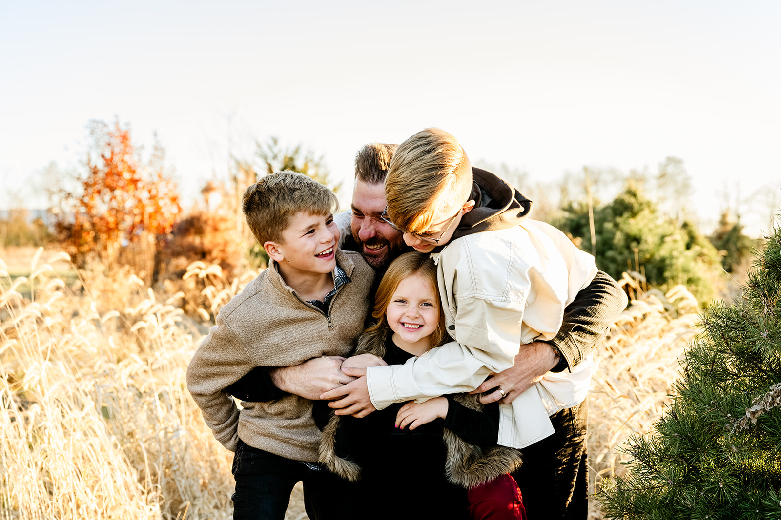 Three happy young siblings jump on and hug dad in a field of tall golden grass at sunset