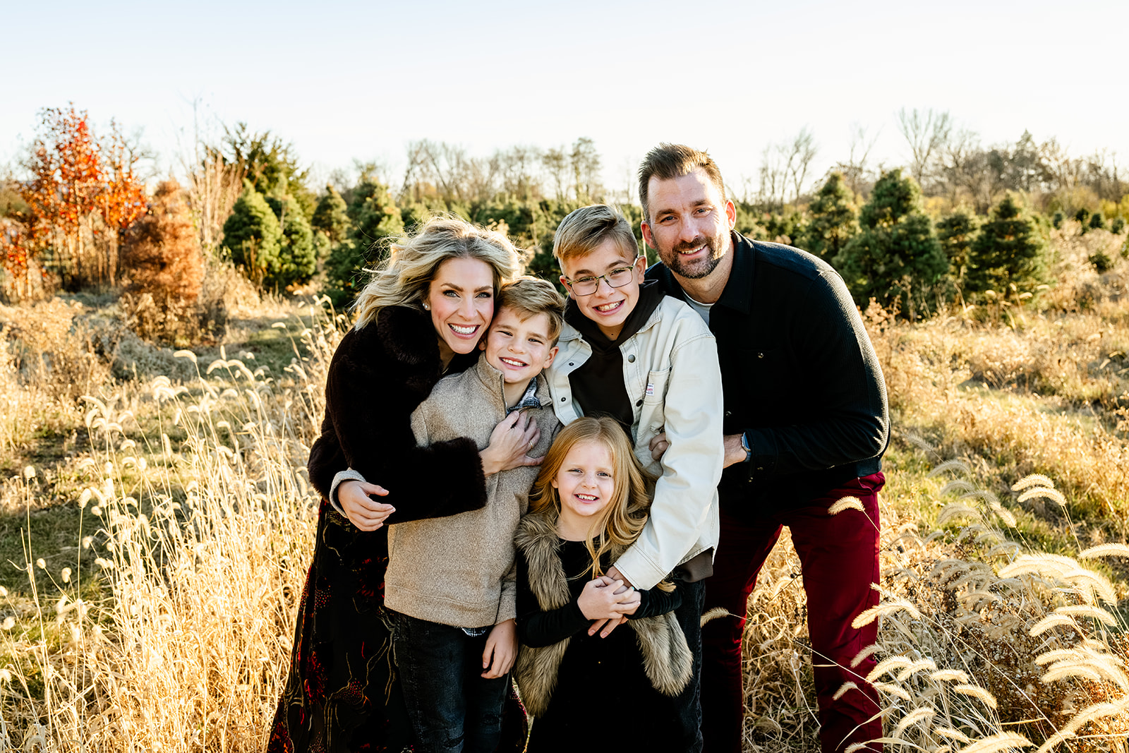 A happy mom and dad smile while hugging their three young children in a christmas tree farm in dark outfits at sunset before finding birthday party places in Naperville