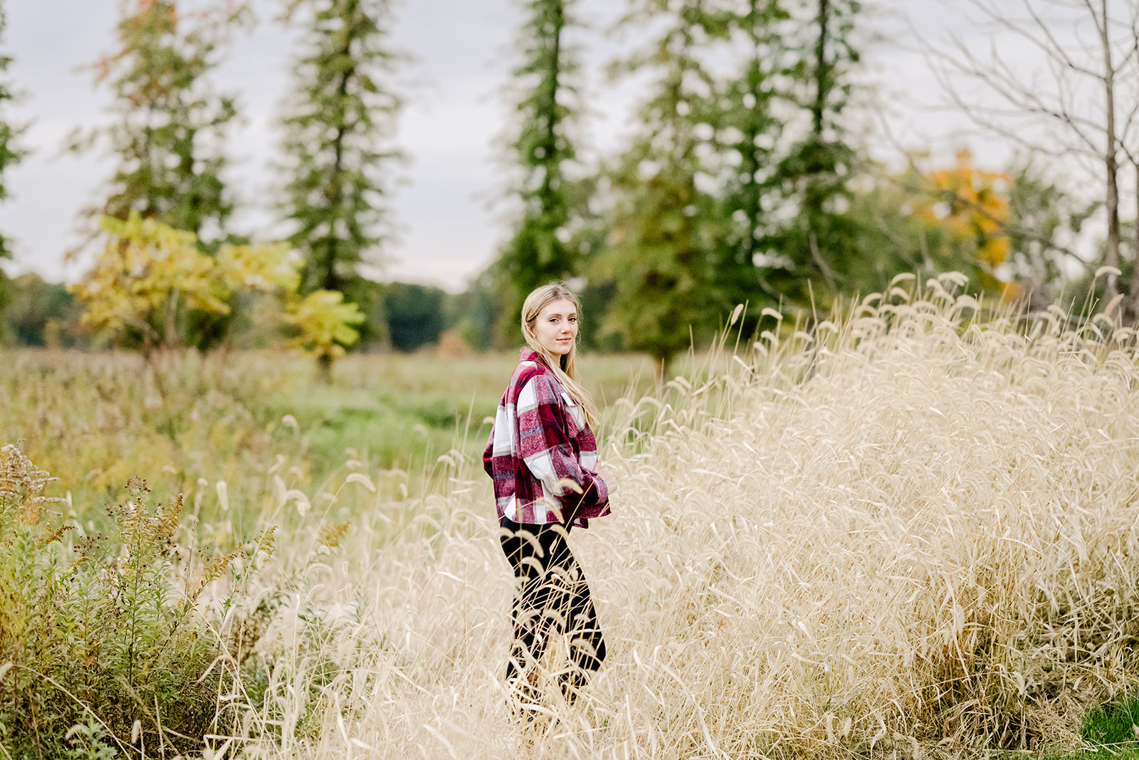 A woman in a red plaid jacket walks in tall golden grasses at sunset and smiling over her shoulder after visiting graduation party venues in Naperville