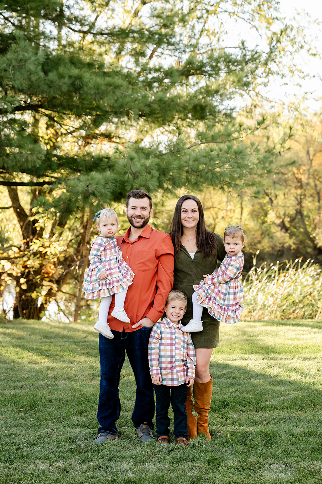 A mom and dad in red and green stand in a park holding two toddler daughters as son hugs mom below them