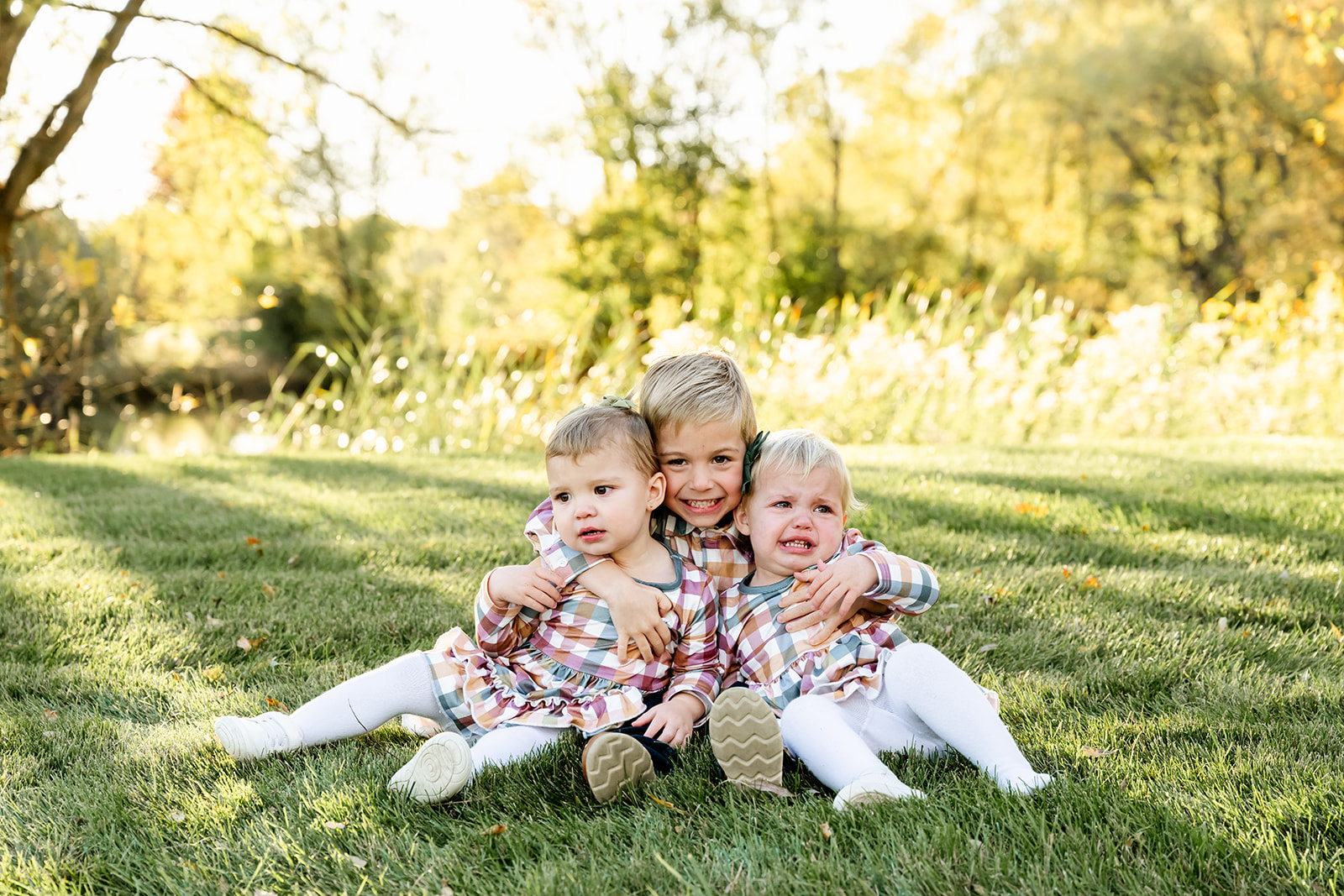 A toddler boy sits in a park lawn at sunset hugging his crying toddler sisters in matching plaid dresses before heading to ice cream shops in Naperville