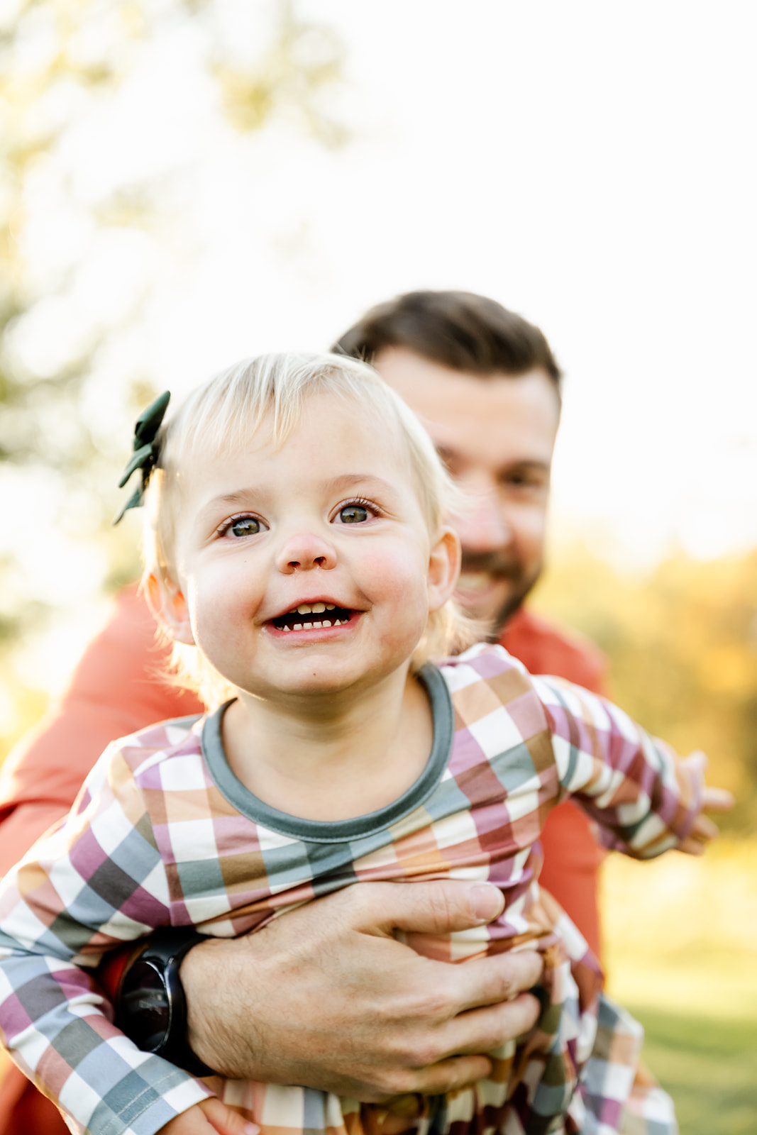A happy toddler girl in a plaid dress is held back by dad while playing in a park at sunset before visiting ice cream shops in Naperville